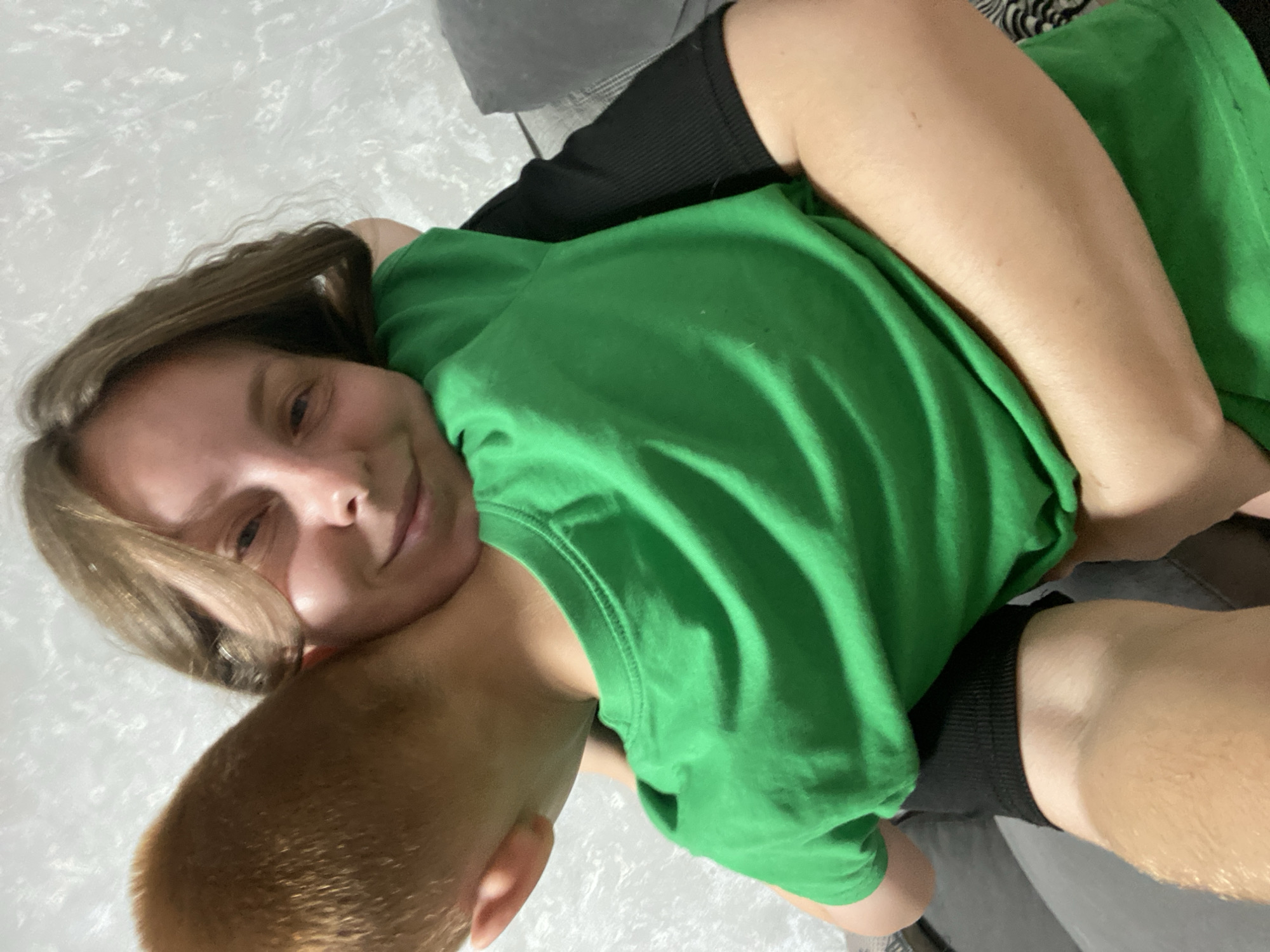 The media file shows two people, a woman and a child, in a close embrace. The woman is lying on her back with the child, who appears to be a young boy, resting on top of her, facing away from the camera. The woman has light brown hair and is wearing a bright green t-shirt and black shorts. Her facial expression suggests a relaxed and affectionate mood. The child has light brown hair and is wearing a black t-shirt.
They are in an indoor setting, possibly a living room or bedroom, as indicated by the presence of furniture, including a gray sofa. The flooring appears to be a light-colored tiled surface. The lighting suggests it could be daytime, with natural light entering the room.
The overall atmosphere of the image is one of warmth and intimacy, capturing a tender moment between a mother and child. There is no visible text in the image. The location is identified as Noviy, Russia.