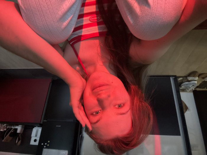 An upside-down view reveals a woman in a striped red and white top and white pants. She is reclined, with her head supported by her hand. The scene is bathed in red light. Behind her, a glossy black surface is visible, possibly a countertop, with some appliances underneath. A glimpse of the floor and some shoes can be seen in the background.