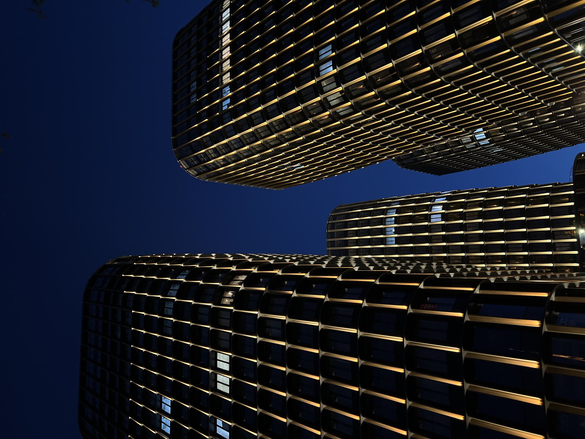 The image captures a low-angle view of three contemporary buildings in Moscow, Russia, at night. The buildings are illuminated with gold trim outlining their dark, rounded facades. The dark blue night sky fills the background. Windows are interspersed throughout the facades with some showing interior lights on. No people are visible in the image.