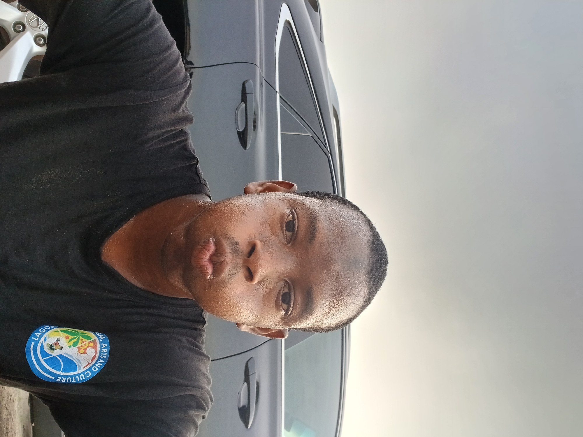 A man with moist skin is in the foreground. He has short, dark hair and is wearing a black T-shirt with a circular logo bearing the words "Lagos," "Arts and Culture," and other imagery. Behind him, a silver or gray vehicle is partially visible, with a window and door handle. The background is a cloudy, light-colored sky. Based on the T-shirt logo, the location is likely somewhere in the city of Lagos, Nigeria. The man is looking at the camera with pursed lips.