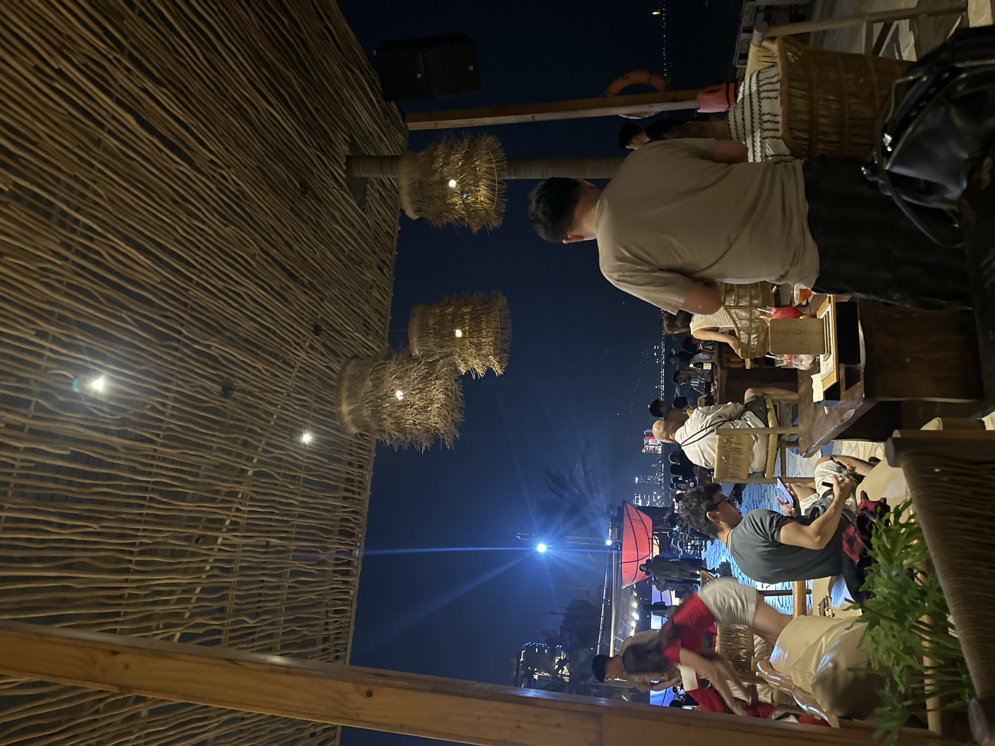 The image depicts an outdoor seating area at a restaurant or bar in Đà Nẵng, Vietnam at night. The seating area is covered by a thatched roof. Several people are seated at tables, some appearing to be dining or socializing. The background reveals a cityscape and a lit-up outdoor stage or event area. The atmosphere is lively, with people gathered together in a public setting.
