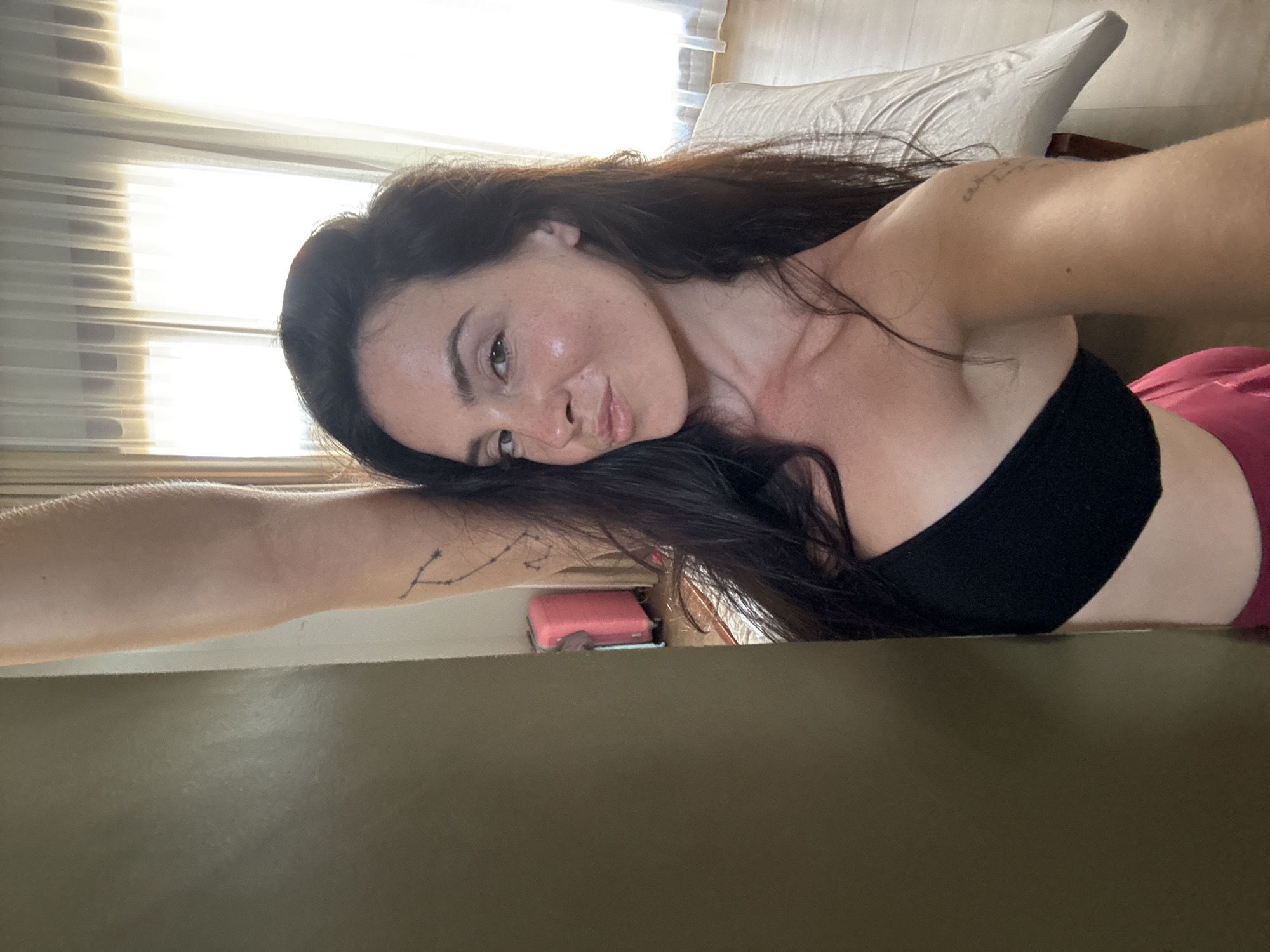 The image shows a woman with long dark hair and light skin, wearing a black strapless top and pink bottoms. She has a tattoo on her upper arm and a constellation tattoo on her left arm. She is standing in front of a wall and window, with a pink suitcase in the background. The room appears to be a hotel room in North Kuta, Indonesia. The woman is looking at the camera and smiling. The lighting is natural and bright.