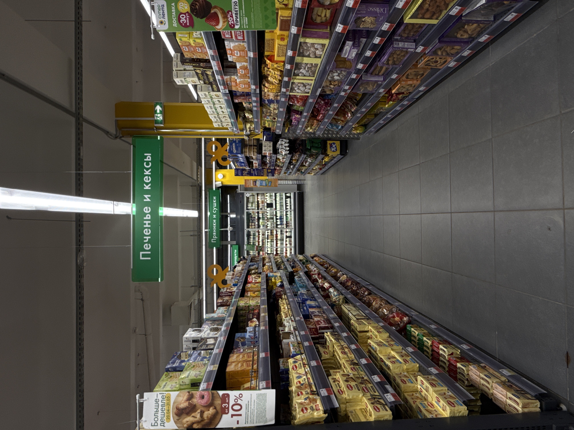 This image depicts a long, well-lit aisle within a Lenta supermarket. The perspective is from the front of the aisle looking towards the back. Two rows of dark-grey metal shelving units line both sides of the aisle, densely stocked with various packaged confectionery items such as cookies, biscuits, crackers, and cakes.
On the left side, a large white promotional sign advertises "More for less!" (Больше-дешевле!) with dates 20.04-31.05 and offers a "10% off from 3 pcs." discount, illustrated with pastries and a donut. Products on this shelf are frequently in yellow packaging.
On the right side, a green promotional sign displays the "Супер Лента" (Super Lenta) logo, advertising "DELIVERY" (ДОСТАВКА) within "30-70 minutes" and a "20% PRIVET20" discount.
Overhead, several green rectangular signs are suspended from the ceiling. The most prominent one in the center reads "Cookies and Cakes" (Печенье и кексы). Further down on the right, another sign indicates "Gingerbread and Sushki" (Пряники и сушки). A smaller sign on the left reads "products" (продукты).
The ceiling features exposed ventilation ducts and multiple linear fluorescent light fixtures, providing bright illumination. The floor is composed of large, dark-grey square tiles. Midway down the aisle on the right, a yellow structural pillar is visible, featuring a green exit sign with a white arrow pointing to the right. In the far background, refrigerated display cases filled with various products can be seen at the end of the aisle. No people are visible in the scene.
This retail environment is identified as a Lenta supermarket in the city of Moscow, Russia, based on the branding and Cyrillic text visible on the signage.
