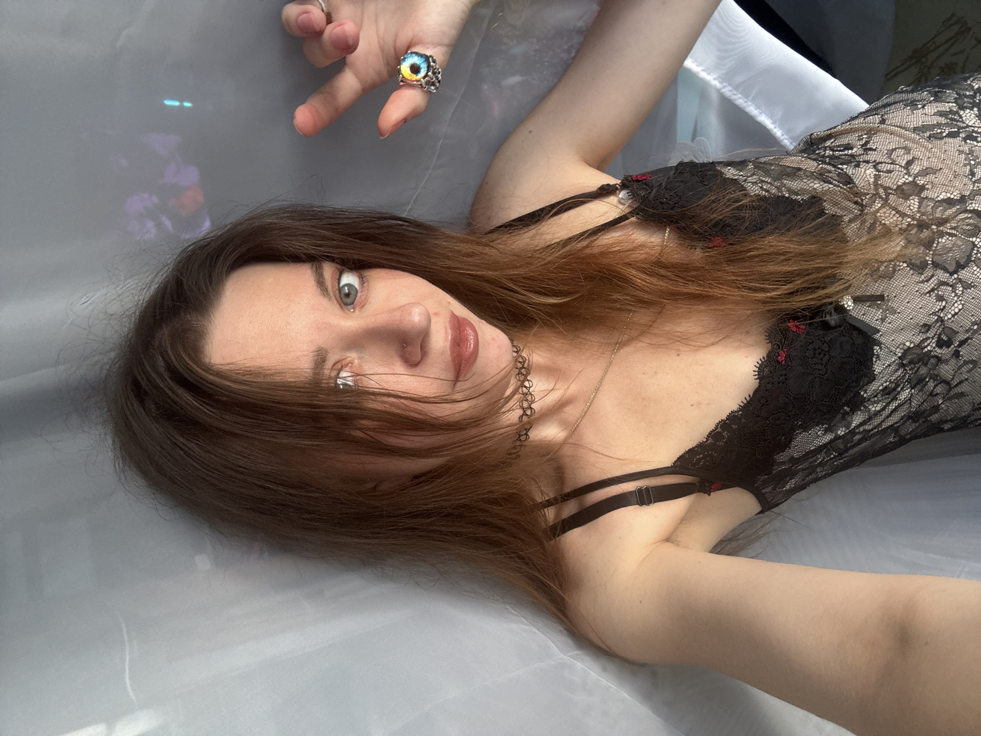 A young woman with long, brown hair is leaning against a sheer, white curtain. She is wearing a black lace bodysuit with red accents. She has a choker necklace and a gold chain necklace. On her left hand, she is wearing a ring with a colorful, eye-shaped stone. The background appears to be a wall with some blurry, colorful lights. The lighting is soft and diffused, casting gentle shadows. The woman is looking directly at the camera with a neutral expression.