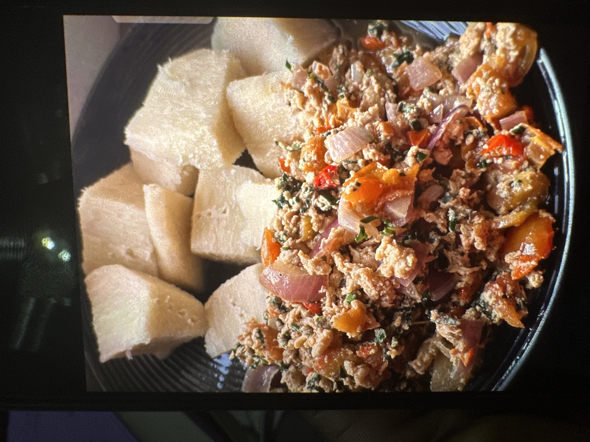 This is a close-up, overhead shot of a black bowl filled with a meal. The bowl is presented on a dark surface, possibly a table, with a hint of a dark screen or frame visible at the bottom and edges, suggesting the image was taken with a phone. The lighting is somewhat diffused, creating soft highlights and shadows.
The meal itself is divided into two distinct parts. On the left side of the bowl are several irregularly shaped pieces of what appears to be boiled yam, cut into bite-sized chunks. They are pale white and have a slightly porous texture.
On the right side of the bowl is a scrambled egg dish. The eggs are cooked and mixed with chopped red onions, red bell peppers, and green herbs, likely parsley or cilantro. The dish has a slightly wet, saucy consistency, with the ingredients distributed throughout the scrambled egg.
The scene depicts a prepared meal, ready to be eaten. There are no people visible, nor any specific time of day or weather cues. The setting appears to be indoors, possibly a home or restaurant. The focus is entirely on the food, emphasizing its textures and colors. The overall impression is of a simple, hearty meal. There is no visible text in the image. The location context of Rido, Nigeria, suggests a potential cultural relevance for this type of dish.