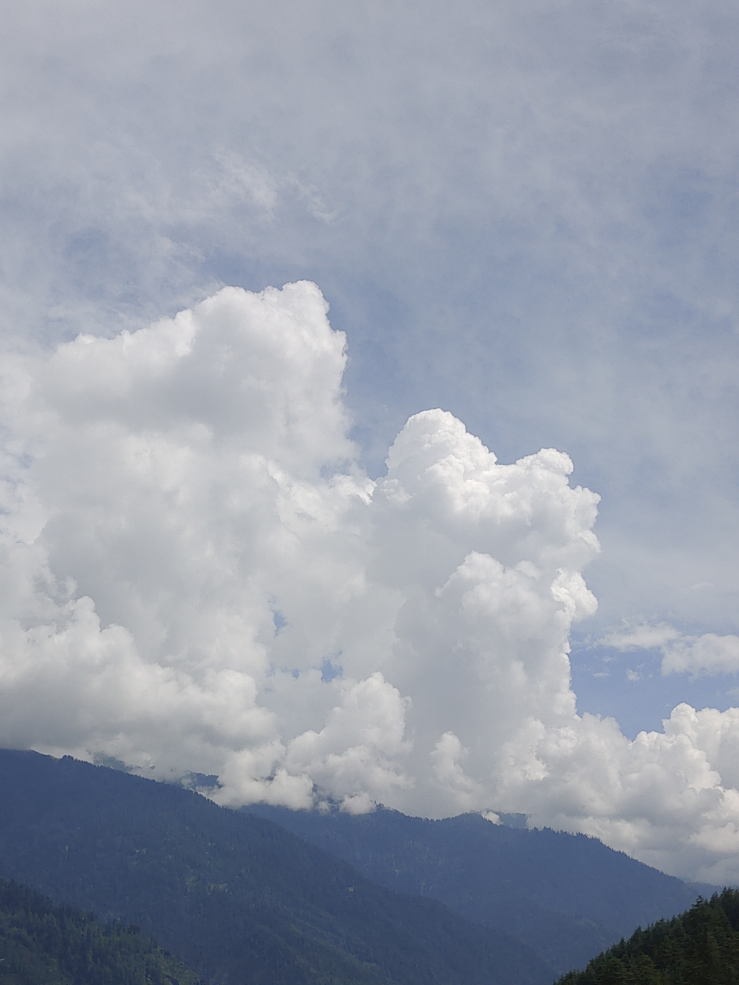 A dramatic sky fills the top two-thirds of the image, featuring a massive cumulus cloud formation in the center, its white peaks stark against a hazy blue sky. The lower third of the image depicts a mountainous landscape, with dark, densely forested slopes receding into the distance. The mountains appear rugged and steep, with their upper ridges partially obscured by lighter, wispy clouds. The overall impression is one of a vast natural landscape under a dynamic sky. This scene is likely from Manali, India, a popular hill station known for its stunning Himalayan vistas.