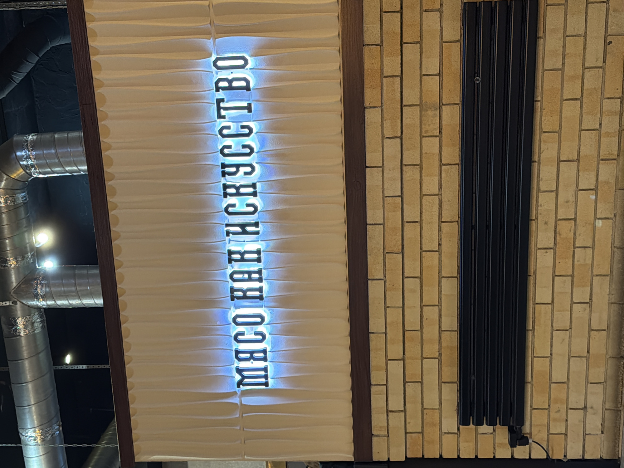 The image shows a sign in Chaikovsky, Russia, which reads "МЯСО КАК ИСКУССТВО" (MEAT AS ART) in vertically stacked, illuminated blue letters. The sign is mounted on a textured white wall, framed by dark brown wood. To the left of the sign, there are cylindrical metal air ducts. To the right, there is a brick wall with a black radiator attached.