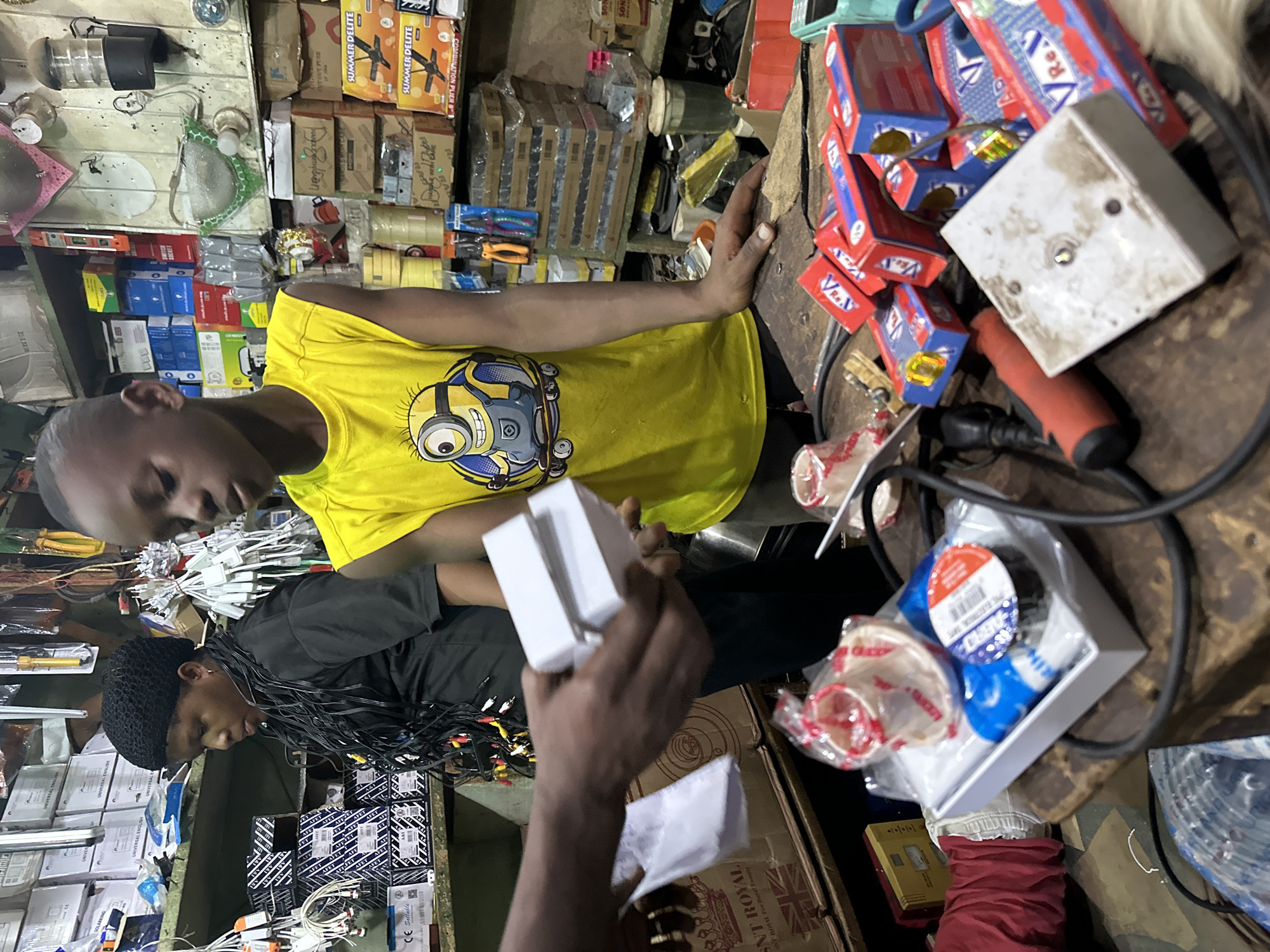 In Barden Yana, Nigeria, a young boy wearing a yellow t-shirt with a Minion graphic stands in a dimly lit, cluttered shop. He appears to be a customer or assistant, holding out a white box in his right hand. A woman in a black t-shirt with a woven hat is visible to his left, her face partially obscured as she looks down. The shop's shelves are filled with various electrical components, boxes, and wires. On a table in the foreground, more electrical supplies are scattered, including small red and blue boxes, a white square object, and electrical tape. The overall impression is a busy, utilitarian retail environment.
