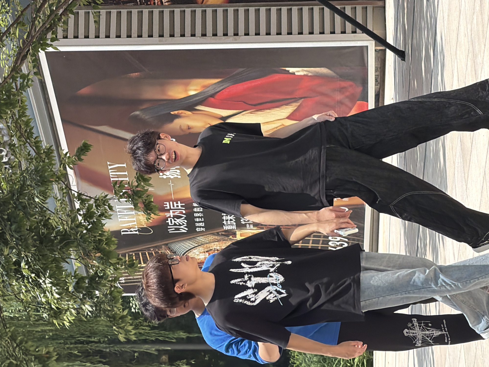 In Yuzhong District, China, three young men walk outdoors in front of a large billboard. The man in the foreground, on the left, wears a black t-shirt with white stylized lettering and light blue jeans. Next to him, the man in the center wears a black t-shirt with a green logo and dark, baggy pants. The third man, to the left and slightly behind, is partially visible wearing a blue t-shirt and black pants with a graphic design. The billboard behind them features a woman in a red jacket. Lush green foliage hangs from trees overhead, casting dappled shadows on the paved ground. The overall scene suggests a bright, sunny day in an urban environment.