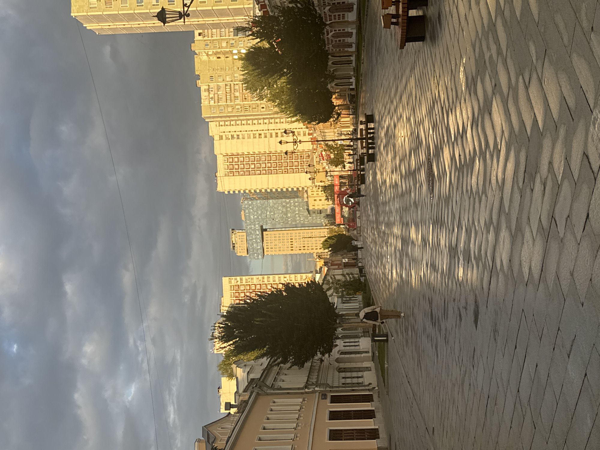 A street in Moscow, Russia, features a person walking down a cobblestone path lined with buildings and trees. The path is wet and reflects the sunlight, creating a shimmering effect. On the left side of the street, there are older, low-rise buildings with arched windows. On the right side, there are taller, modern apartment buildings. Trees are planted along the sidewalks. In the distance, more modern buildings, including a glass skyscraper, are visible. The sky is cloudy, with patches of sunlight breaking through. The scene appears to be captured during the golden hour, with the low sun casting long shadows. There are streetlights and benches along the path.