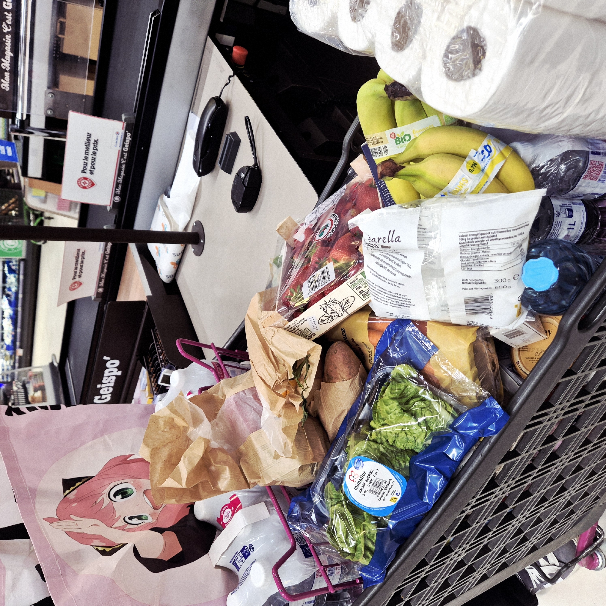 The image shows a shopping cart filled with groceries at a checkout counter in a store, likely in Geispolsheim, France, based on the "Geispo" branding. The cart contains items such as toilet paper, bananas labeled "BIO Villa," strawberries, a bag of "farella," lettuce in a blue package labeled "mimaflor Salade Sucrine," bottled water, and various packaged goods in brown paper bags. A pink shopping bag with an anime character is visible on the side of the cart. The checkout counter has a payment terminal, a scanner, and signage promoting "Mon Magasin C'est Geispo'" ("My Store is Geispo'").