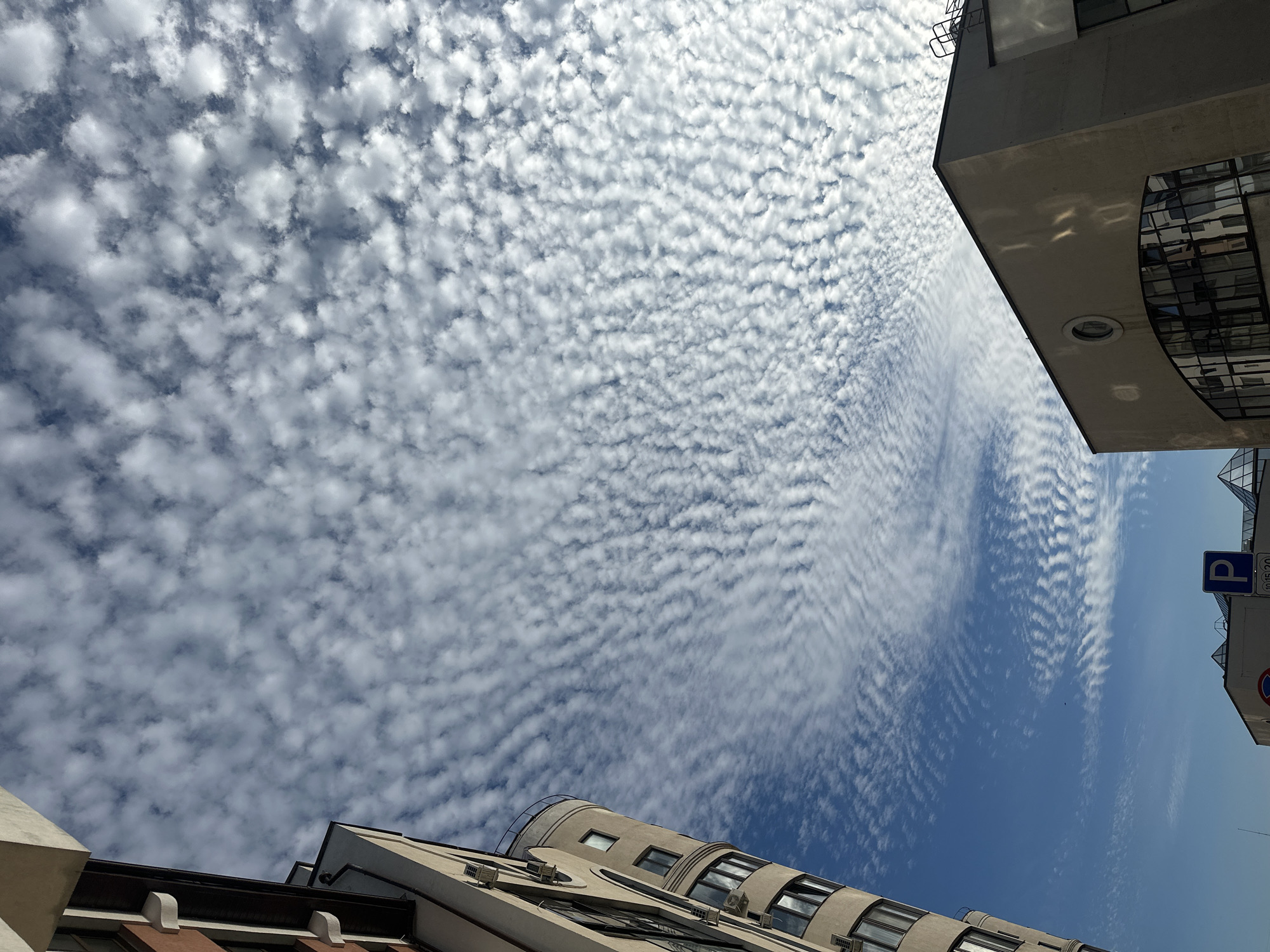 The image shows a cityscape with buildings and a sky full of altocumulus clouds. A tall, modern building with rounded sections and multiple windows is visible on the left. Another building with a flat roof and large arched windows is on the right. In the lower part of the image, there are parking signs, including a blue sign with a white 'P'. The sky is a clear blue with a dense pattern of small, white, puffy clouds that create a ripple-like effect. The scene appears to be captured from a low angle, looking upwards towards the sky. The location is specified as Ufa, Russia.