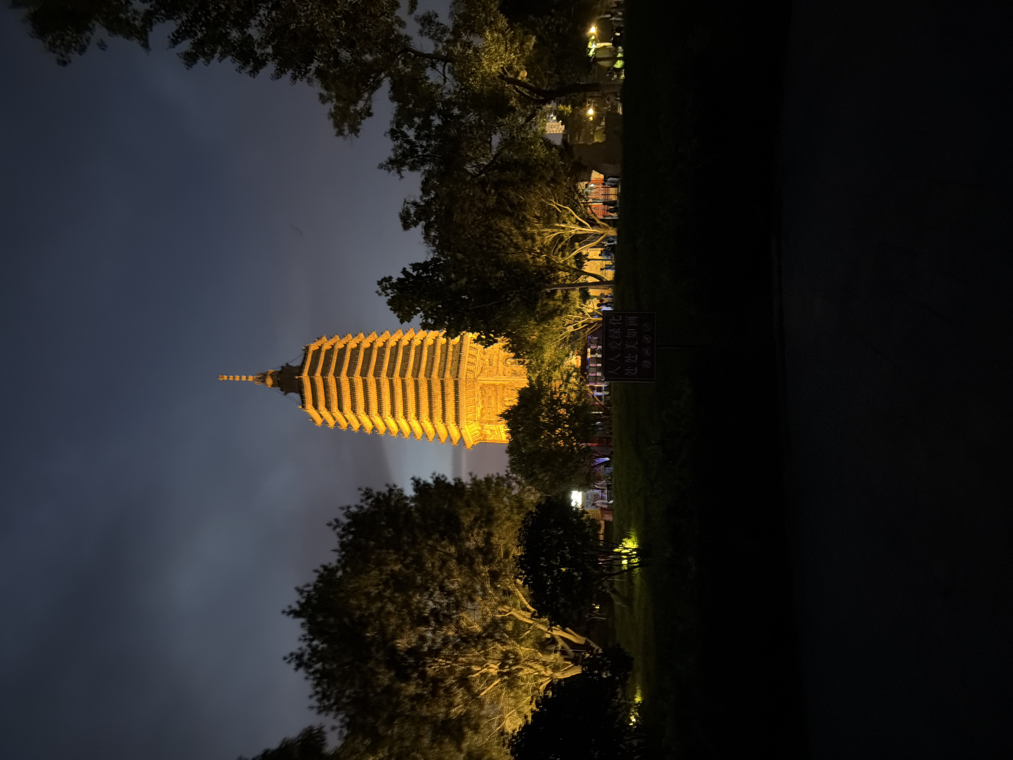 The image captures the Jinzhou Tower in Jinzhou, China, at night. The tower, illuminated with a warm golden light, stands prominently in the center of the frame, its multiple tiers reaching towards the dark, cloudy sky. Trees with illuminated foliage surround the base of the tower, creating a park-like setting. In the foreground, a dark path or road is visible, with a signpost containing Chinese characters placed on a grassy area. Several figures are present near the base of the tower, suggesting it is a public space frequented by people in the evening. The overall atmosphere is serene and somewhat mystical due to the dramatic lighting and twilight setting.