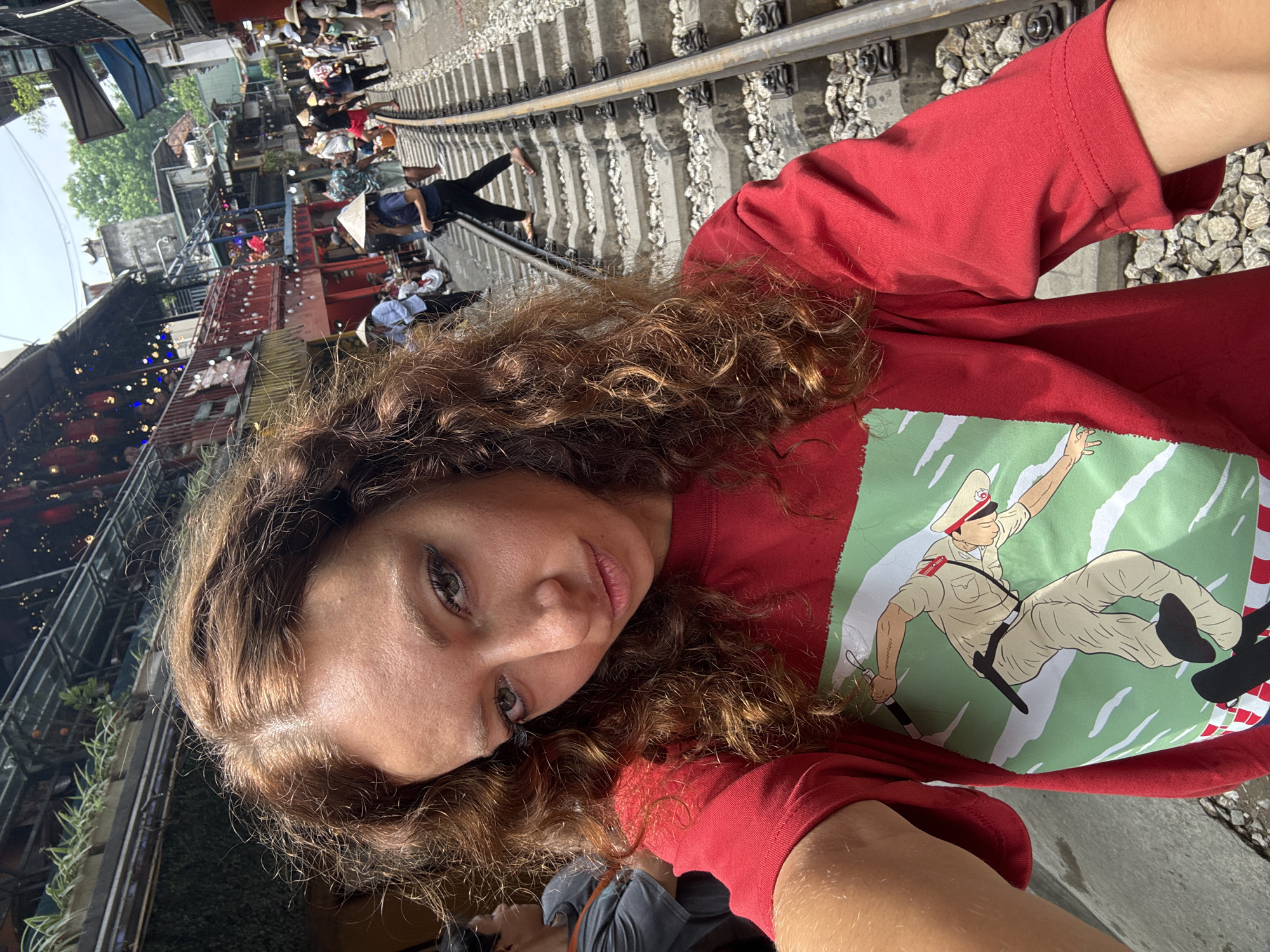 A woman in a red t-shirt with a graphic is taking a selfie on the Train Street in Hà Nội, Vietnam. The train tracks run diagonally across the image, lined with gravel. Tourists are walking along the tracks, some wearing conical hats. Buildings and cafes are visible alongside the railway, with lights strung up. The woman has curly brown hair and is looking at the camera.