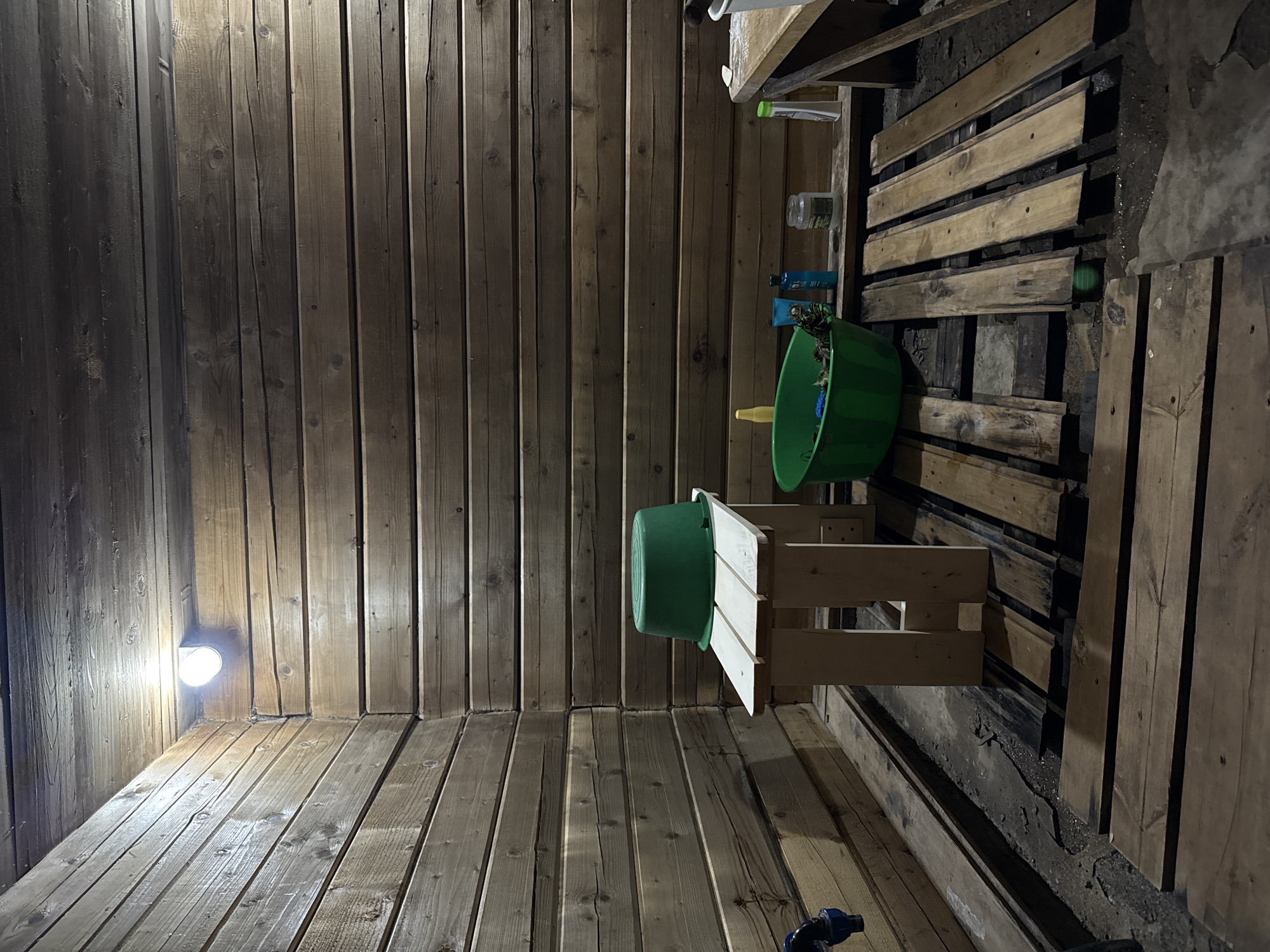 The image depicts the interior of a wooden shed, possibly a potting shed or storage area, in an unknown location, not necessarily in Sludka, Russia. The walls, floor, and ceiling are constructed of vertical wooden planks. A battery powered light is mounted in the corner of the room. A small wooden table or stand holds a green plastic tub or bucket. Another larger green plastic tub is placed above, along with several bottles on a shelf. A wooden pallet leans against the right side. The area appears cluttered but organized.
