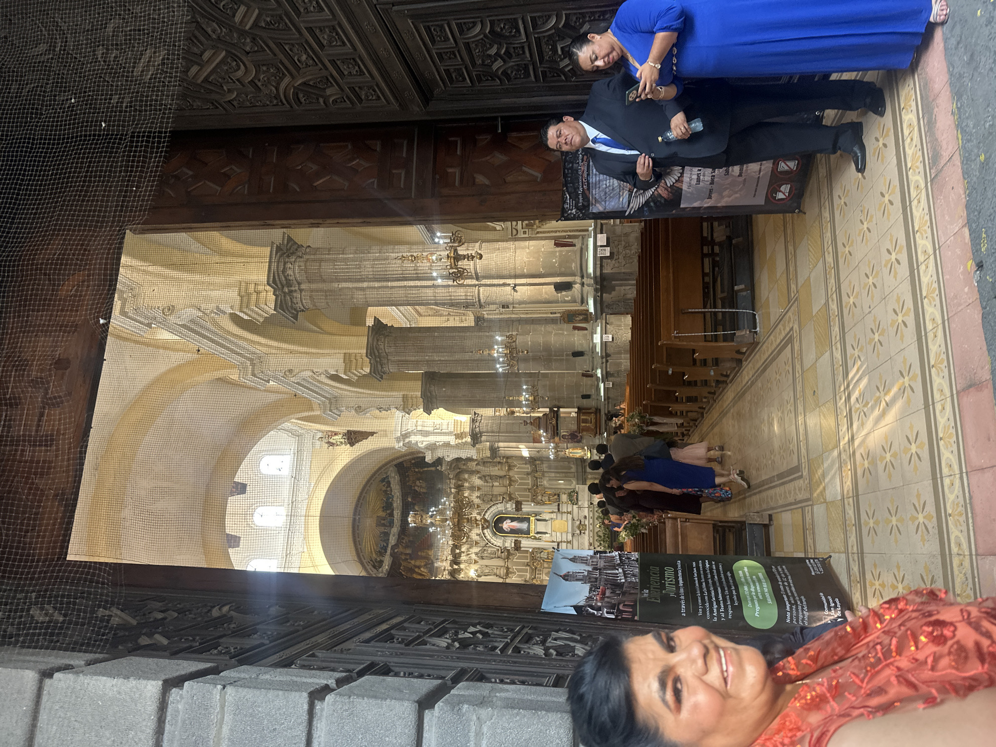 Here is a concise description of the image:
The image captures a partial view of the interior of a church in Puebla, Mexico. Visible is the ornately decorated altar, arches, and pillars typical of religious architecture. Several people are seen inside the church, while a man in a suit and a woman in a blue dress stand near a doorway. A sign indicating the "Temple Expiatorio del Espíritu Santo" is also visible. A woman in an orange dress is at the extreme lower right.