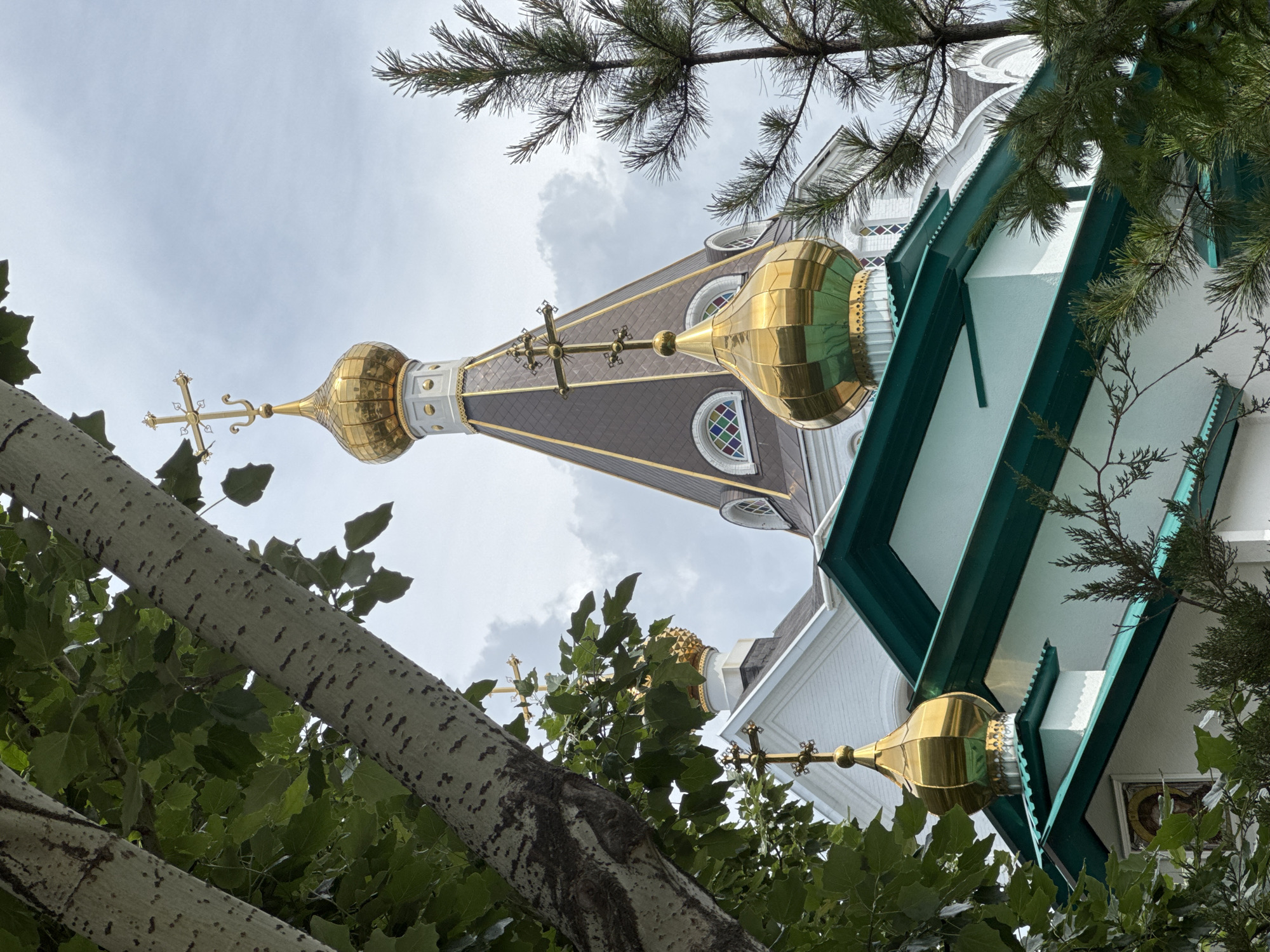 The image depicts a building with a golden dome and cross, characteristic of Russian Orthodox church architecture, viewed from a low angle through the foliage of trees. The building's exterior is primarily white with green trim. The sky is overcast. The location is not discernible from the provided image.