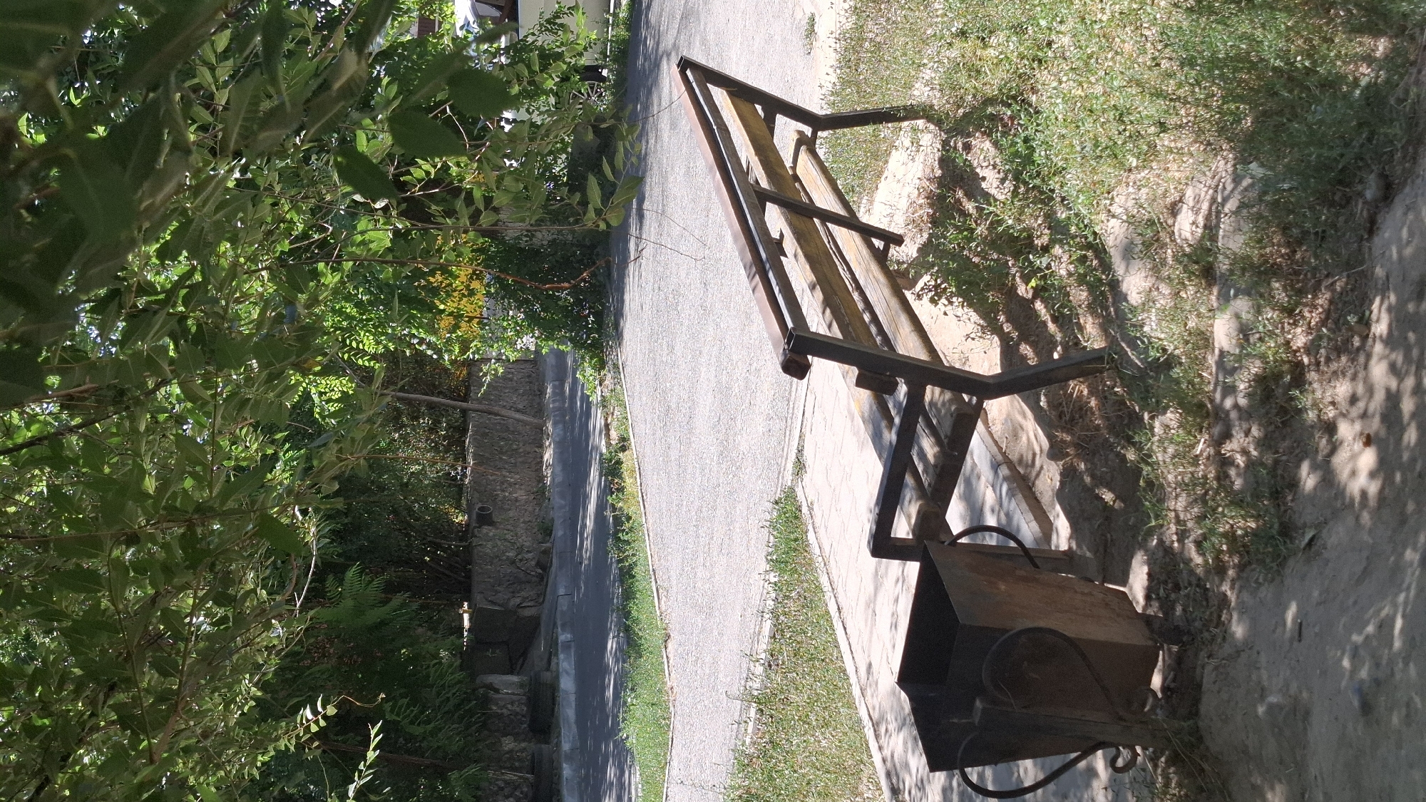 An outdoor scene in Shymkent, Kazakhstan, features a weathered metal and wood bench positioned on a concrete pathway. To the left of the bench, a rusty metal trash receptacle with decorative handles stands on the ground. The scene is shaded by lush green foliage from an overhead tree, casting dappled shadows on the pathway and surrounding dry grass. In the background, a paved road runs alongside a low stone wall. The overall impression is one of a quiet, possibly neglected, public space.
