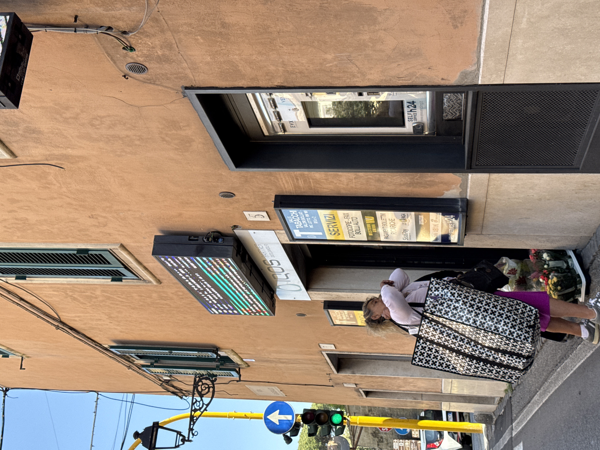 This daylight image, taken in Castel Gandolfo, Italy, captures a street scene with a woman standing near a commercial building. The woman, with blond hair, is dressed in a light-colored top and a purple skirt, holding a large black and white patterned tote bag and a smaller black handbag. She appears to be leaning against the building or resting, looking downwards.
The building features a light brown/tan facade with several signs and windows. A prominent digital sign displays colorful, illuminated text and patterns. Below it, a sign partially reads "CASTEL GANDOLFO" and "SOOCHI". Another large sign, labeled "SERVIZI" (Services), lists offerings such as "TABACCHI" (Tobacco), "FOTOCOPIE - FAX", "BOLLI AUTO" (Car Stamps), "WESTERN" (likely Western Union), "PAGAMENTI BOLLETTINI" (Bill Payments), "RICARICHE" (Recharges), "SisalPay", "LS PAGA", and the "amazon" logo. One window has green louvered shutters. A self-service machine, labeled "SELF SERVICE H24", is visible within an entryway, showing text like "www.ervedding.it", "EVX", and "VENE". Near the woman, some flower arrangements are placed by the entrance.
The scene is brightly lit by the sun, indicated by strong shadows and a clear blue sky visible at the bottom. Street elements include a yellow pole, a blue arrow sign pointing left, and a traffic light displaying a green signal. Various cables are affixed to the building's exterior.