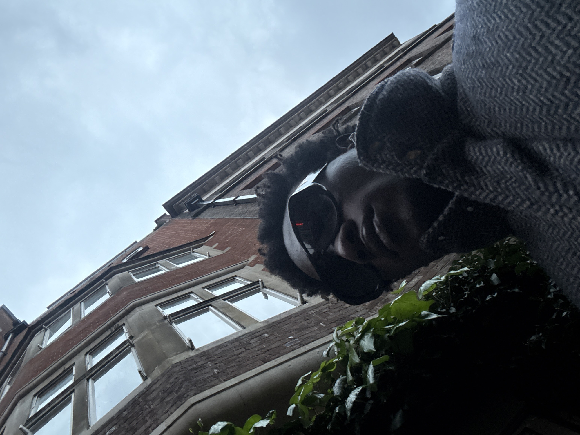 A low-angle shot features a person in the foreground, wearing what appears to be headphones or a headset with a visible red light. The person has dark hair. They also appear to be wearing a coat with a herringbone pattern. The background shows a building constructed of red brick with multiple windows. The sky is overcast and gray. Greenery, potentially ivy, is visible at the bottom right corner of the image. The style of the building suggests a location in London, United Kingdom.