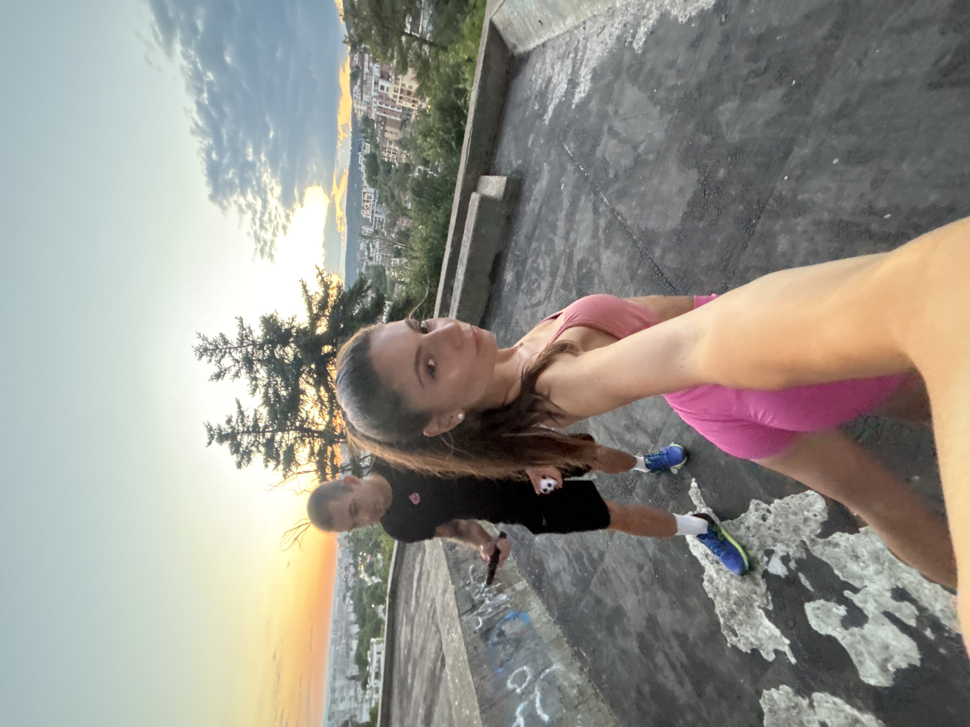 This is a selfie-style photo taken outdoors at sunset, likely from a high vantage point overlooking the city of Varna, Bulgaria. The foreground features a woman in a pink athletic top and shorts, with her arm extended to capture the photo. She has brown hair pulled back in a ponytail and is wearing pearl earrings. Behind her, a man in a black t-shirt and shorts stands with a dark object, possibly a phone, in his hand. He is wearing blue and neon green athletic shoes with white socks. The background shows a city skyline with buildings and trees, illuminated by the warm colors of the setting sun. The surface beneath them appears to be a concrete rooftop or observation deck with some graffiti and patches of what looks like snow or debris. Two evergreen trees are visible to the left of the frame. The sky is a blend of light blue and orange hues, with some clouds present.