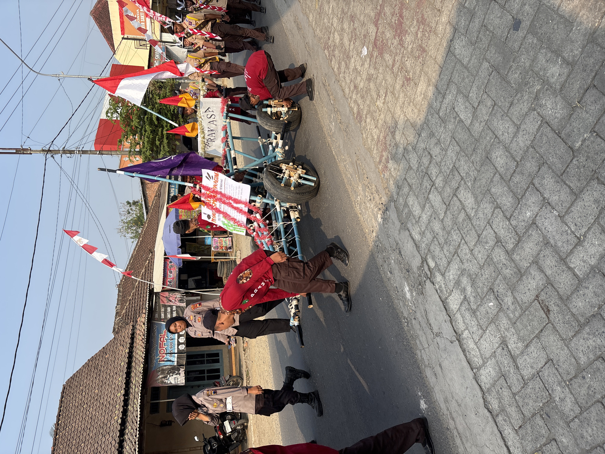 This image captures a street scene in Tirtomoyo, Indonesia, likely during a parade or event. In the foreground, a blue and white contraption resembling a cart or float is being pushed along a paved road. Several individuals are associated with the cart, including a person in a red shirt bending down to adjust one of the wheels, and others in scout uniforms. Behind them, a few people in police uniforms are also present. The background features buildings with tiled roofs and banners, one of which displays text in Indonesian. Several Indonesian flags (red and white) are visible, both on the cart and held by people in the background, along with a purple flag. The overall impression is of a community gathering or procession on a sunny day.