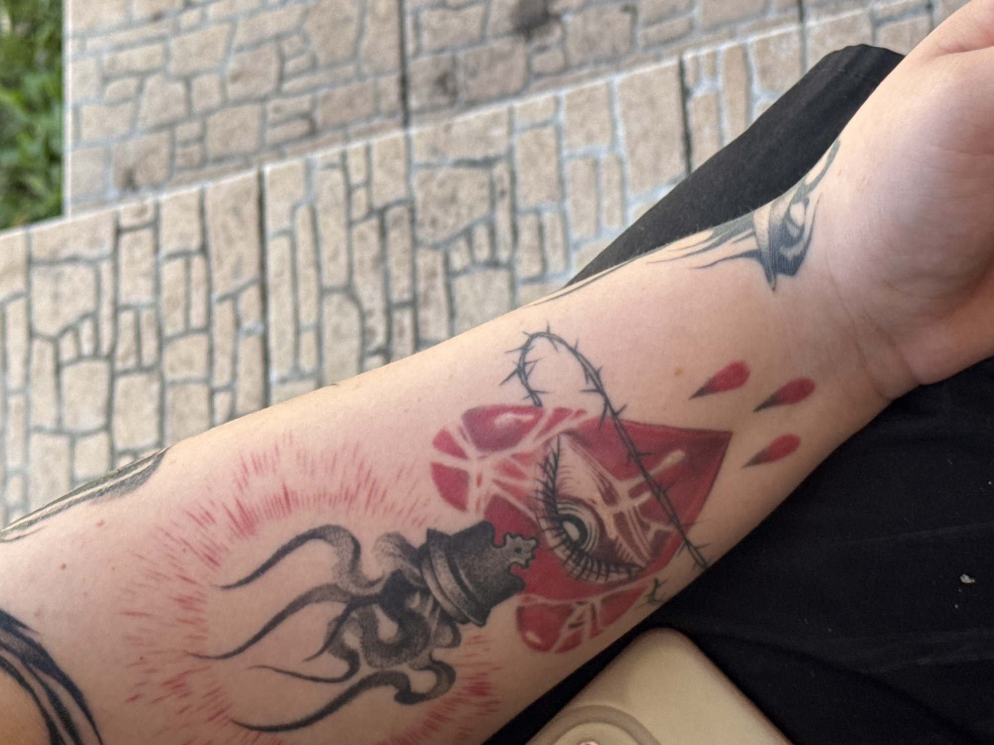 The image shows a close-up of a person's forearm with several tattoos. The tattoos are predominantly black and red and appear to be done in a surreal or symbolic style.
The main subject of the tattoo is a red heart-shaped object with an eye in the center. Barbed wire is depicted wrapping around this heart. To the left of the heart is a black chess piece, a pawn, with swirling, flame-like lines emanating from its base, appearing to be part of another tattoo or design. Around the pawn, there are red lines radiating outwards, resembling light or energy. Three red teardrop shapes are visible to the right of the heart. Further down the forearm, a black tattoo with sharp, angular lines is visible.
The person's skin shows some redness, potentially indicating fresh ink or irritation. The background of the image shows light-colored, rectangular stone blocks, possibly part of an outdoor staircase or wall, with some blurred greenery visible in the upper left corner. The lighting suggests it is daytime. The overall mood conveyed by the tattoos is dark and possibly intense due to the imagery of the eye, barbed wire, and blood-like drops. There is no visible text in the image.