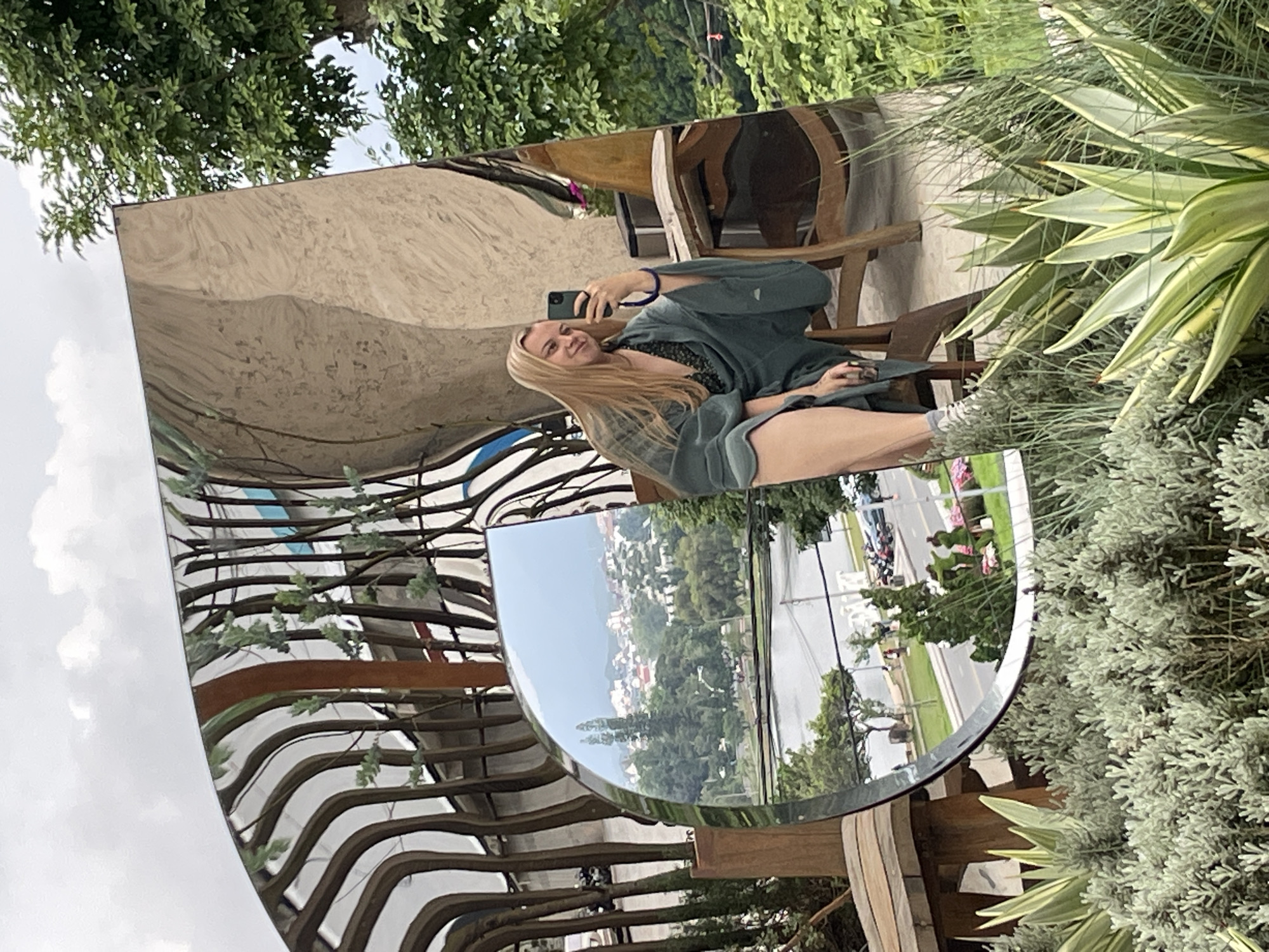 A woman with blonde hair is taking a selfie in a large, distorted mirror in what appears to be an outdoor seating area. She is wearing a dark patterned top and a teal kimono-like robe, sitting on a wooden chair. The mirror reflects a panoramic view of a city with white buildings and red roofs nestled amongst lush green hills, a body of water, and trees. The foreground of the image shows various plants, including succulents and bushes. The background features a canopy of trees. The overall setting suggests a cafe or restaurant with a scenic view in Da Lat, Vietnam, known for its hilly terrain and French colonial architecture, which aligns with the reflected cityscape.