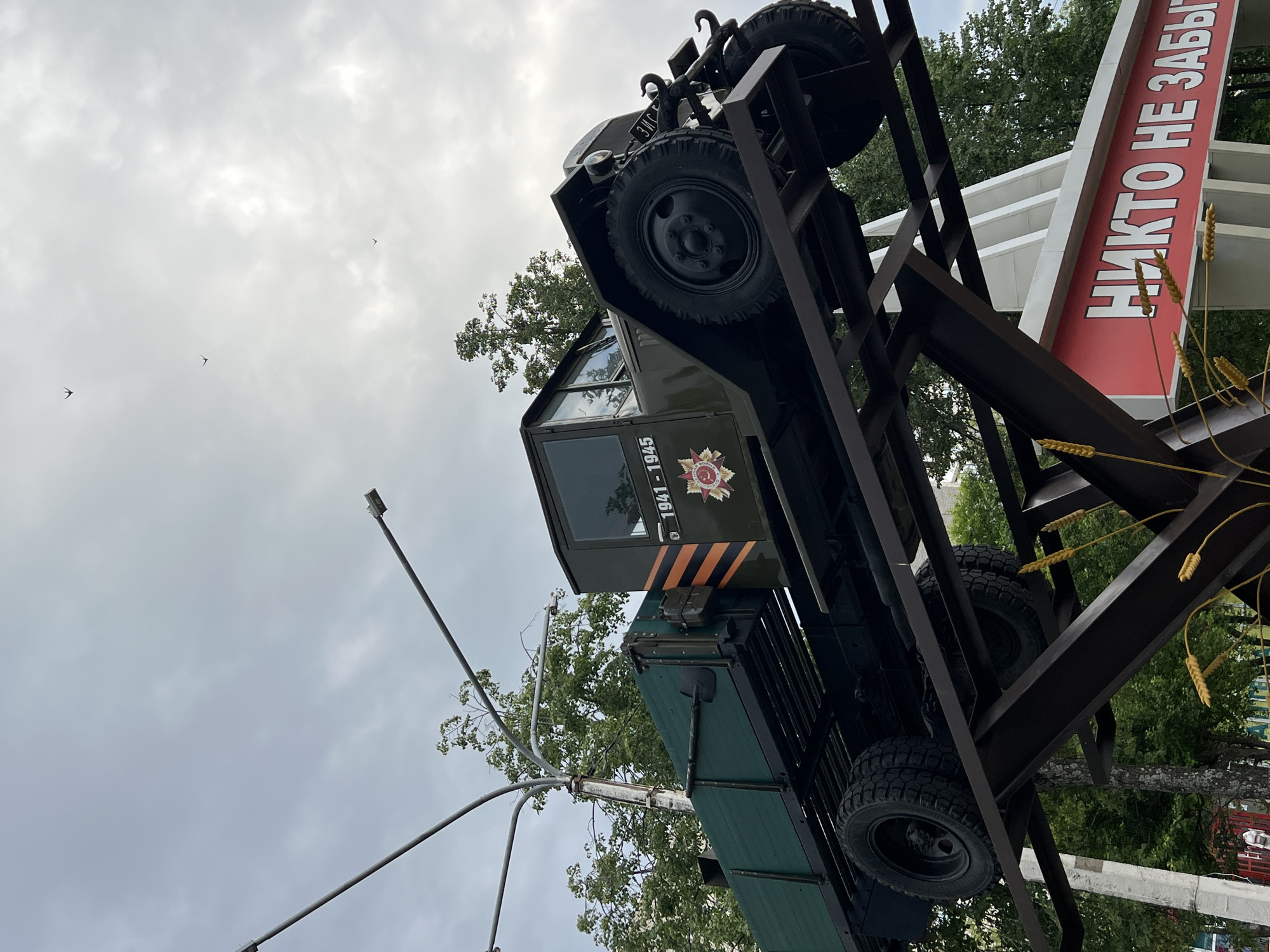 An olive green vintage Soviet military truck, likely from World War II, is displayed on a metal stand, elevated above street level. The truck has the years "1941 - 1945" and a red star emblem with "СЛАВА" (Glory) in Cyrillic script on its side. It is positioned in Chaikovsky, Russia, as indicated by a red sign below with white Cyrillic lettering that reads "НИКТО НЕ ЗАБЫТ" (No one is forgotten). The truck is parked at an angle, with its front facing towards the right of the frame. Behind the truck, trees and a cloudy sky are visible. Several birds are flying in the sky. The overall scene suggests a war memorial or historical exhibit.