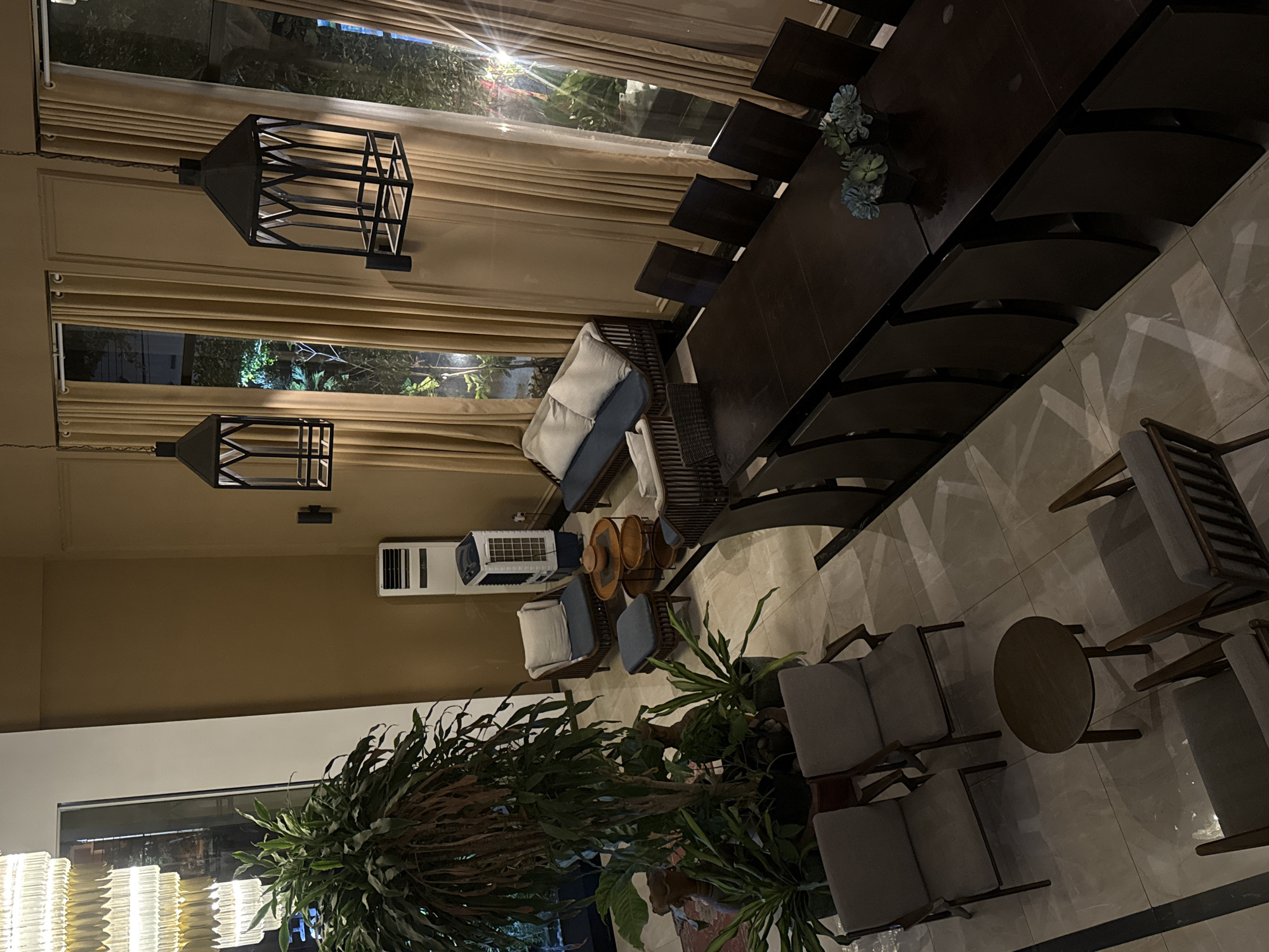 An indoor scene, possibly a hotel lobby or restaurant, is depicted from a high-angle perspective. The space features a large dining table with multiple chairs, a seating area with sofas and armchairs, and decorative elements like plants and lighting fixtures. Two large, caged pendant lights hang from the ceiling, casting shadows. Tall curtains cover large windows, revealing a dimly lit exterior. The flooring is tiled. The overall atmosphere is subdued, with warm lighting and a focus on furniture and decor. The location is identified as Bac Nha Trang Ward, Vietnam. No individuals are visible in the image.