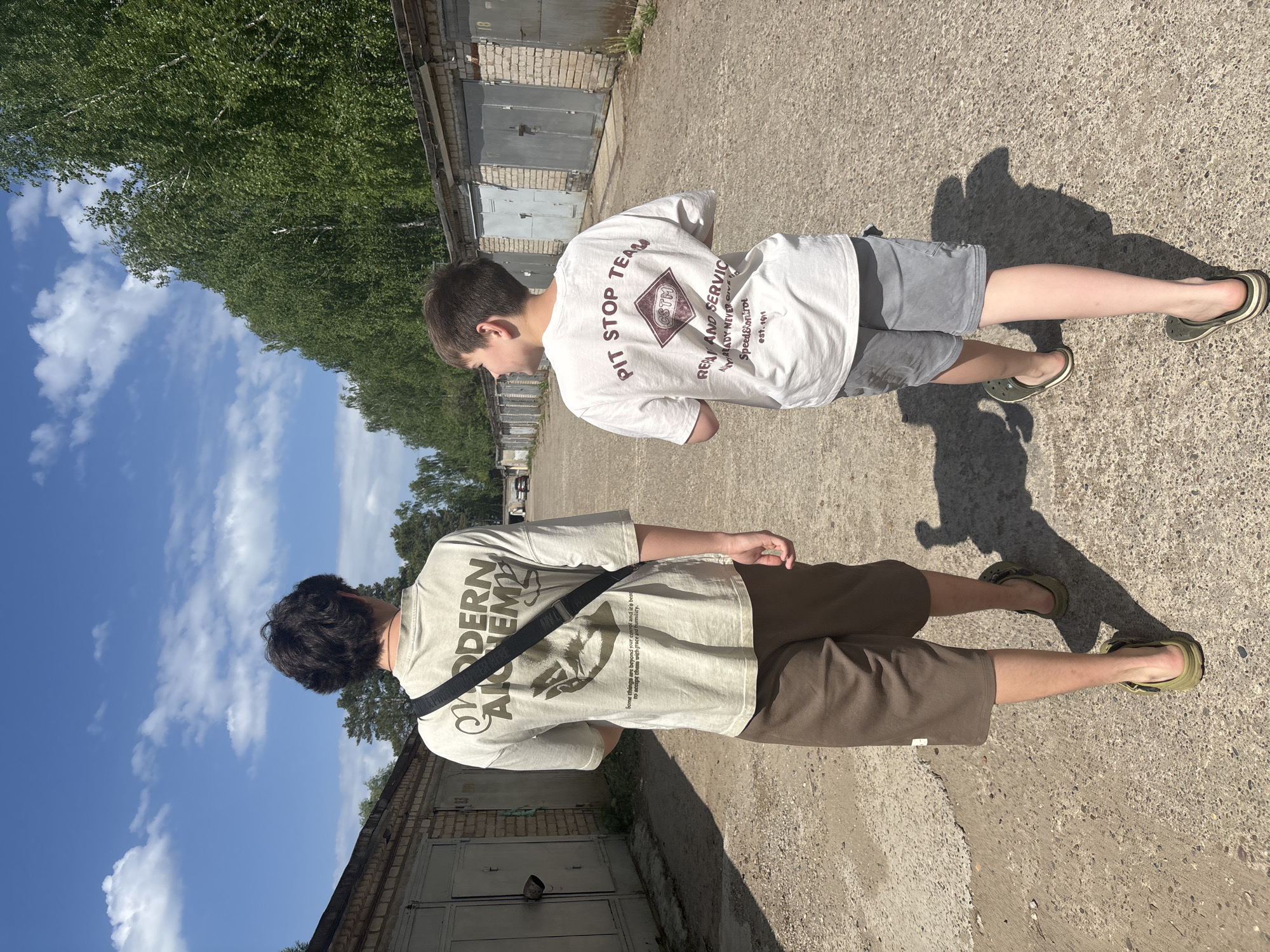 Two youths stand in a narrow alleyway between garage doors, possibly in Chaikovsky, Russia. One wears a light brown "Modern Alchemy" t-shirt with a black strap across the chest, along with brown shorts and green sandals. The other wears a white "Pit Stop Team" t-shirt, grey shorts, and green sandals. The alley is paved and partly shaded by foliage from nearby trees. The sky is visible with scattered clouds. The garage doors are old and weathered.