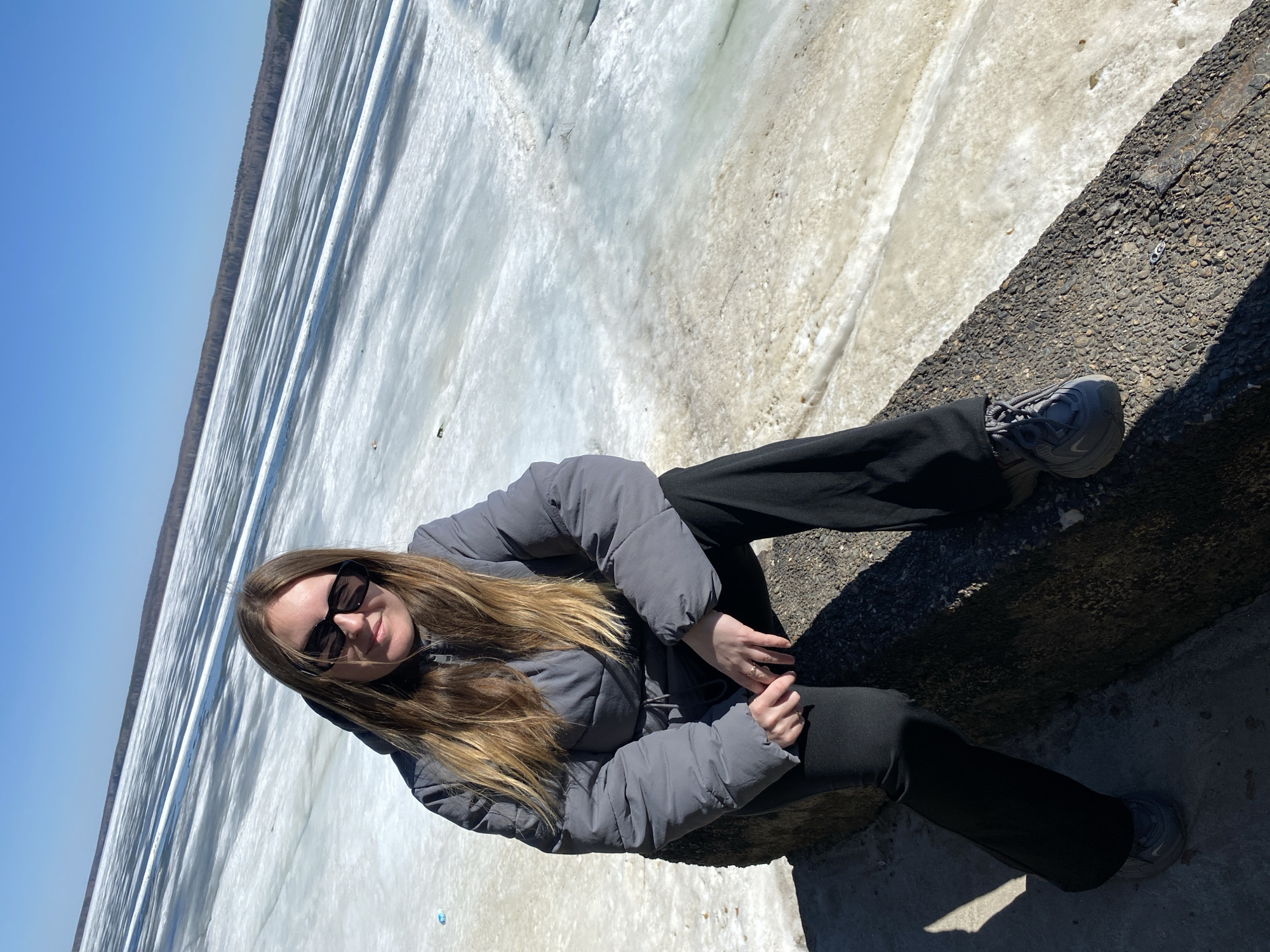 A young woman wearing a gray puffer jacket, black pants, sunglasses, and gray sneakers is sitting on a concrete structure adjacent to a frozen body of water, possibly a river or lake, on a sunny day in Chaikovsky, Russia. The ice cover appears partially melted in areas. The sky is a clear blue. A line of trees can be seen along the distant shore.