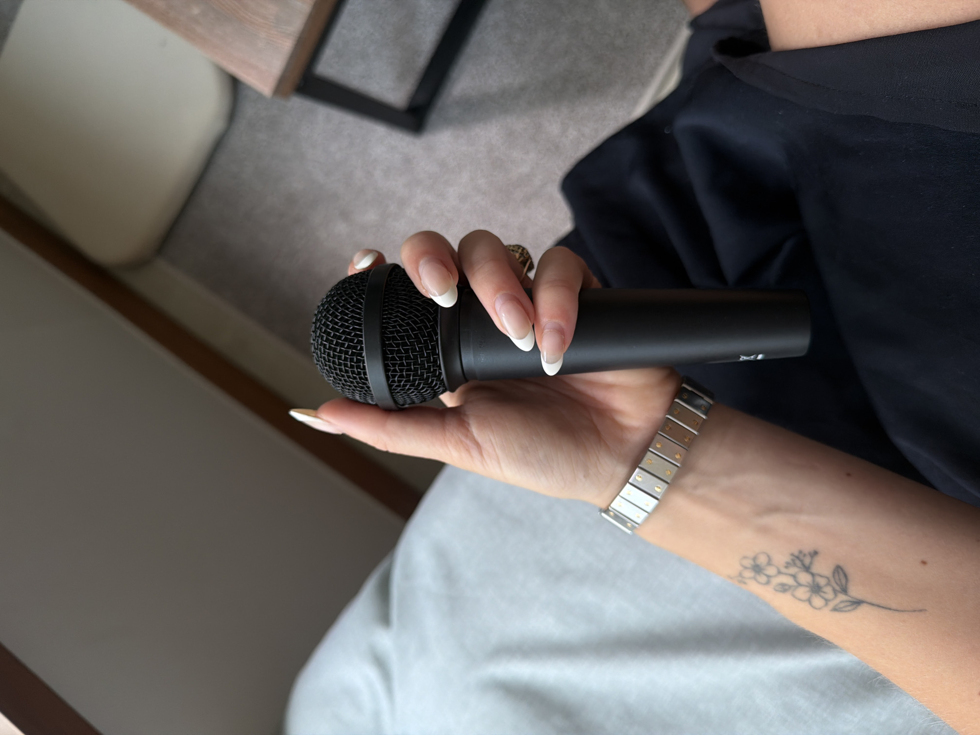 A person with manicured nails holds a black microphone in their right hand. The person wears a silver and gold bracelet on their right wrist, and a small floral tattoo is visible on their forearm. The background includes a portion of a wooden-framed table and neutral-toned soft furnishings. No information in the picture indicates the location as Seoul, South Korea.