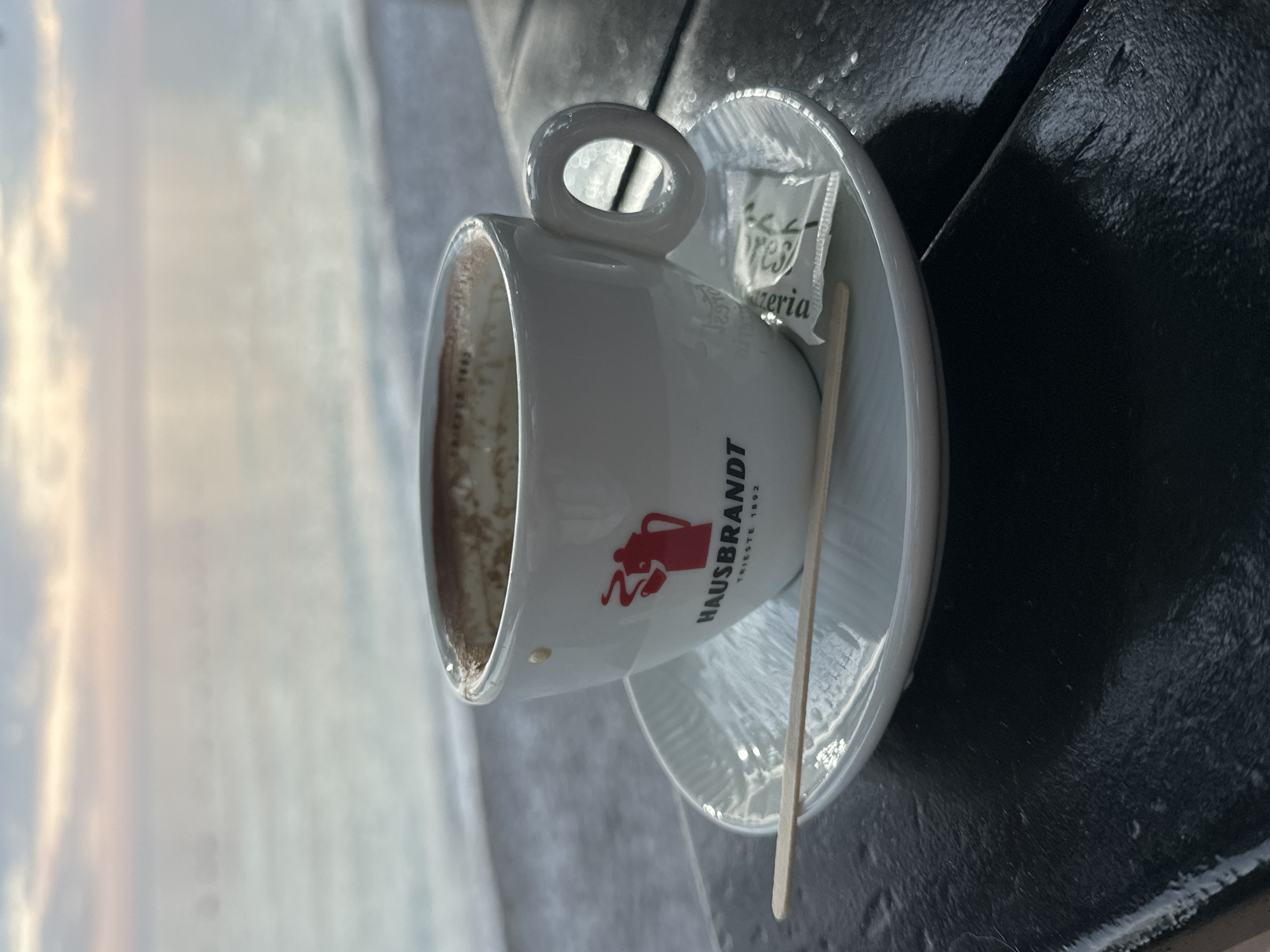 A close-up, slightly tilted shot shows a white ceramic coffee cup with the red "HAUSBRANDT TRIESTE 1892" logo resting on a matching saucer. The cup is nearly empty, with traces of coffee residue visible. A wooden stirrer and a small sugar packet are placed on the saucer. The cup and saucer sit on a dark, possibly weathered, surface. The background reveals a blurry coastal scene with the sea and sky suggesting either sunrise or sunset. This scene is identifiable as Bar, Montenegro due to the Hausbrandt logo, a coffee brand widely available in that region, and the coastal setting characteristic of Bar.