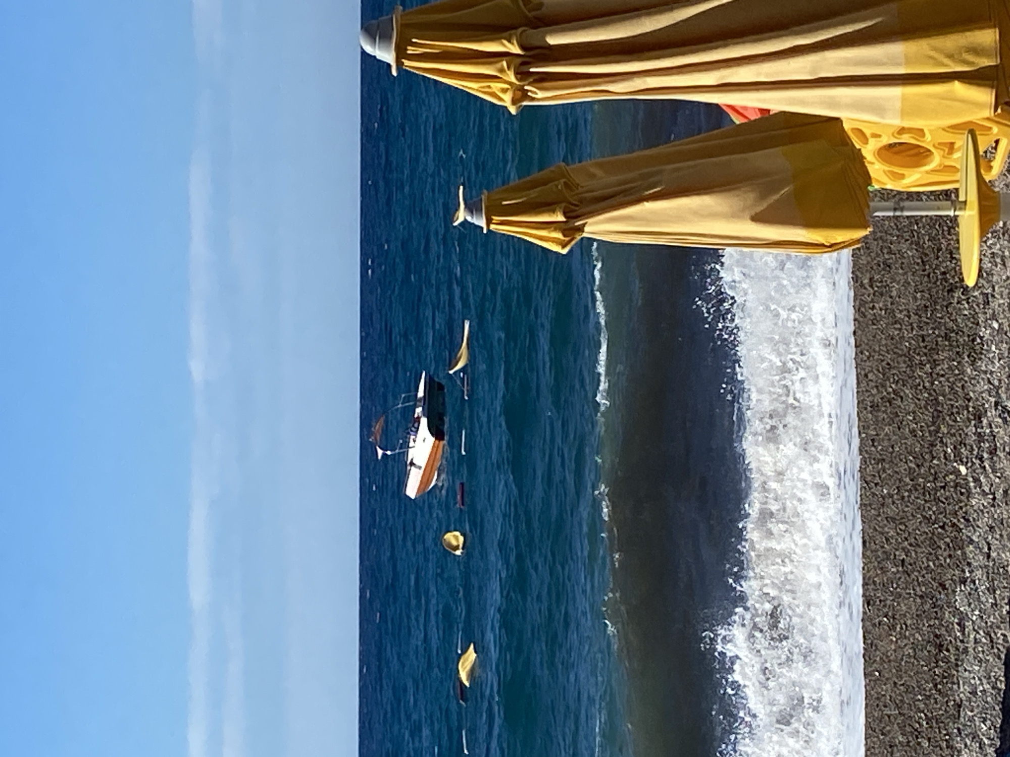 The image depicts a bright, sunny day at a beach in Prijevor, Montenegro. The foreground is dominated by two closed, yellow beach umbrellas and a small yellow table. The beach itself is covered in dark pebbles. Beyond the shore, the deep blue sea is active with waves breaking, creating white foam. In the water, a small white and orange motorboat is visible, along with several yellow buoys or floats. The sky is a clear, light blue with a few wispy clouds. The overall scene conveys a serene, yet lively, beach atmosphere.