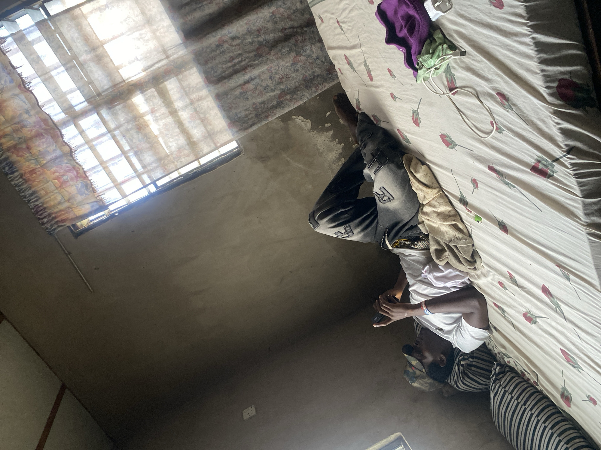 The image shows two people in what appears to be a bedroom. One person is lying on a bed, facing away from the camera. They are wearing a white t-shirt, dark pants, and a patterned head covering. Their right arm is bent, and they are holding a dark object, possibly a phone, in their hand. A light-colored cloth is draped over their legs.
Another person is sitting on the edge of the bed, their back mostly to the camera. They are wearing a dark and white striped shirt and dark pants. Their head is angled downwards, and they are looking at something in their hands, which are not clearly visible.
The bed has a white sheet with a red rose pattern. There are also a few other items on the bed, including a purple garment and a green cloth. The room has simple walls and a window with blinds or curtains. Sunlight streams through the window, creating bright patches on the wall and bed. The overall lighting suggests it is daytime. The scene appears casual and domestic.