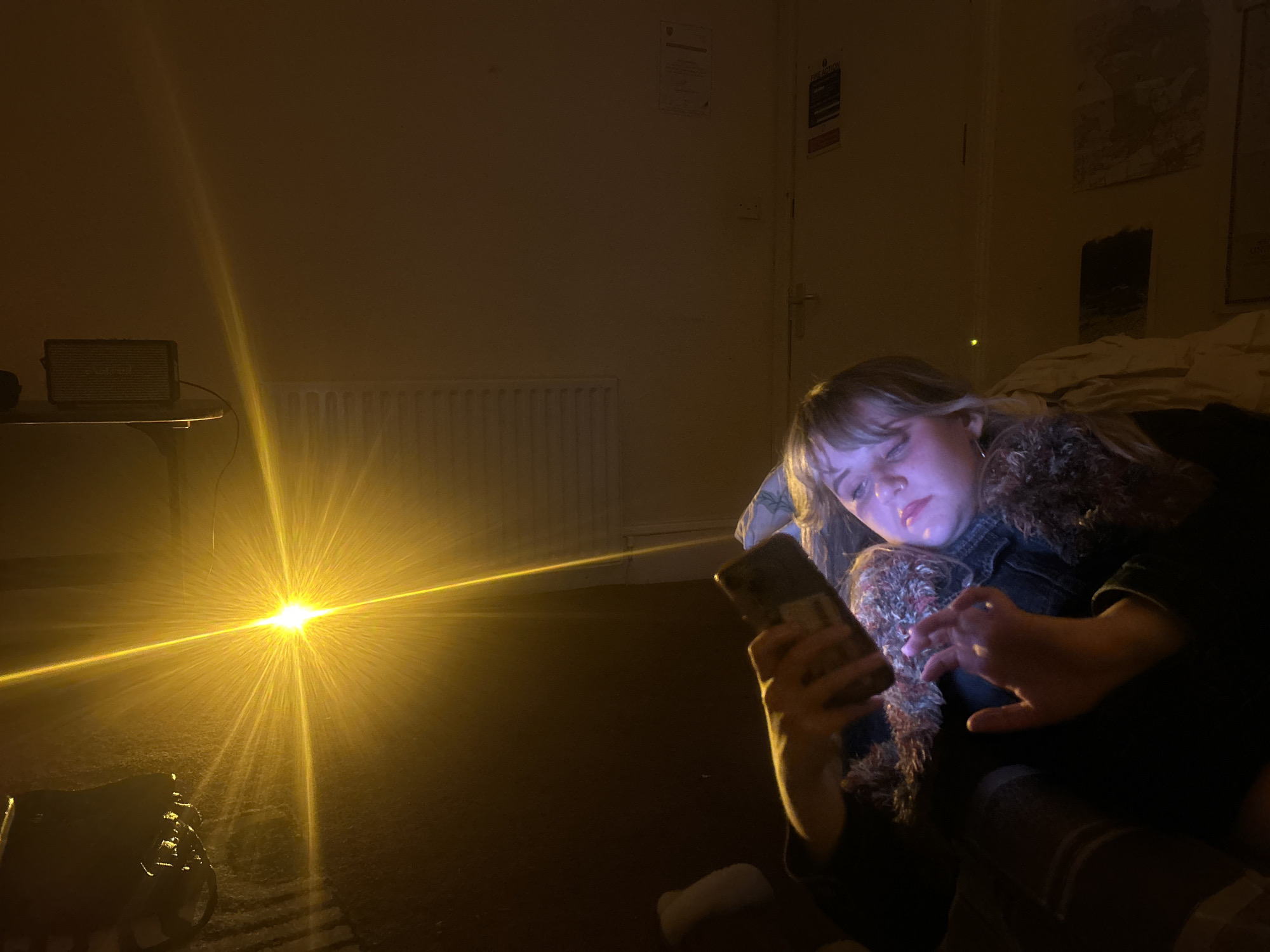 This image depicts an indoor scene, likely a bedroom, in a dimly lit environment, suggesting nighttime or a low-light setting. A young person, appearing female with light-colored hair and bangs, is positioned on the right side of the frame, reclining or lying down. They are engrossed in a smartphone, which emits a distinct blue light that illuminates their face. They are wearing what appears to be a denim jacket with a fluffy, possibly faux fur, collar or trim.
A very bright, yellow light source with prominent lens flares and streaks emanates from the lower-left side of the image, dominating that half of the frame. On a table in the mid-left, a rectangular electronic device, resembling a speaker (possibly Marshall branded), is visible with a cable connected. Below the table, a white, horizontally ribbed radiator is mounted on the wall.
In the background on the right, a closed, light-colored door is visible. Above the door and to its right, several documents or posters are affixed to the wall. One clearly visible sign is a blue and white "FIRE ACTION" notice. The room appears to have a carpeted floor. The overall atmosphere is one of quiet solitude or relaxation. This scene is located in a room in Dundee, United Kingdom.