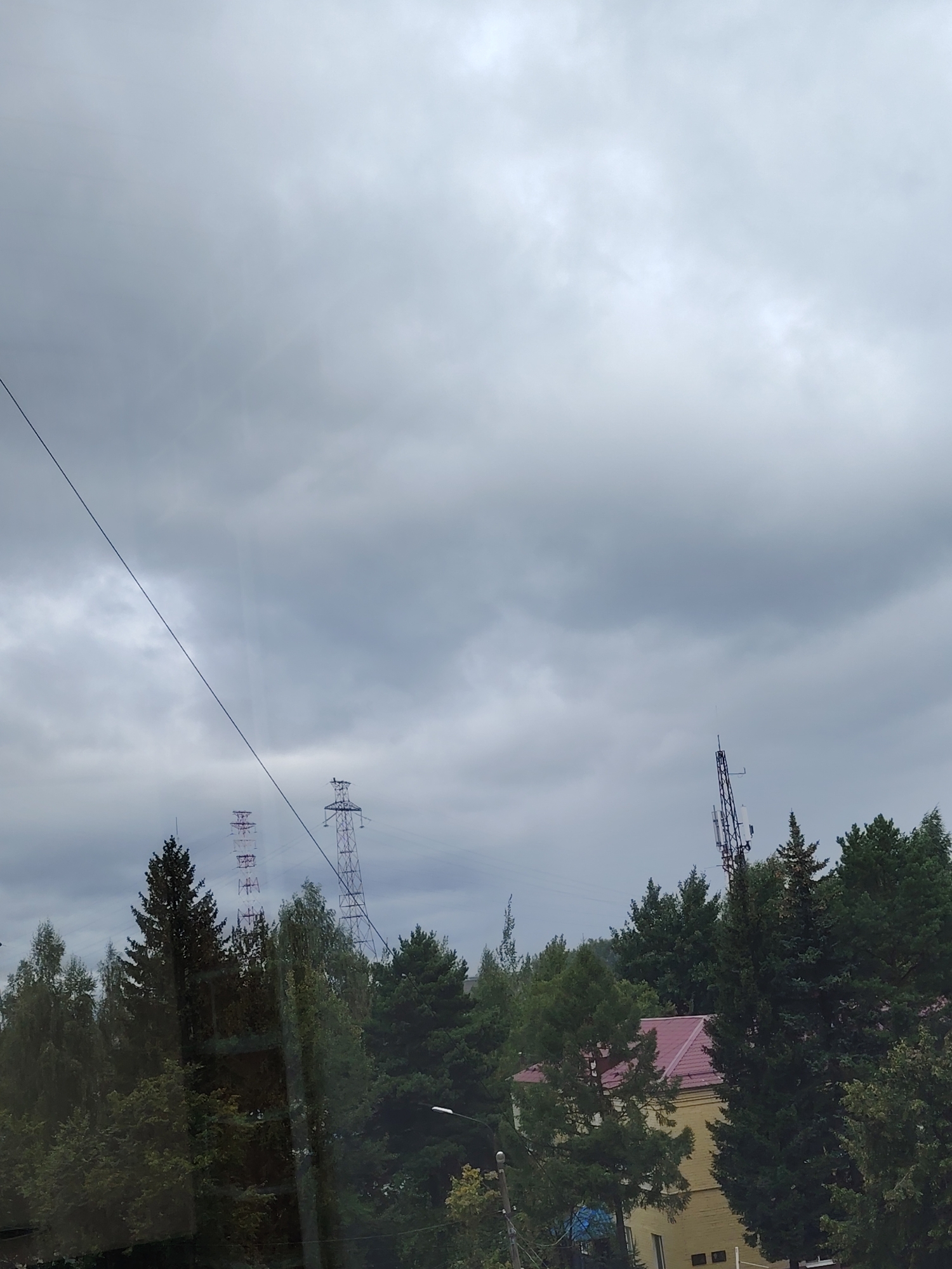 The image shows an overcast sky with scattered clouds, casting a gray, diffused light. In the foreground, a dark, thin wire stretches diagonally across the frame from the upper left to the lower middle. Below the wire, a dense line of trees with varying shades of green foliage stretches across the lower half of the image. These trees appear to be mostly coniferous, with some deciduous trees interspersed.
Rising above the trees, several structures are visible. To the left, two tall, lattice-work electricity pylons are partially obscured by the trees. The pylon on the left has some red accents. To the right of these, a more slender, dark metal tower, possibly a telecommunications mast, stands prominently, topped with an antenna. Behind the trees and to the right of the mast, a portion of a building with a red, pitched roof and yellow walls is visible. The scene suggests a semi-rural or suburban setting on the outskirts of Konakovo, Russia, given the presence of electricity infrastructure and a mix of natural and built environments. No people are visible in the frame.