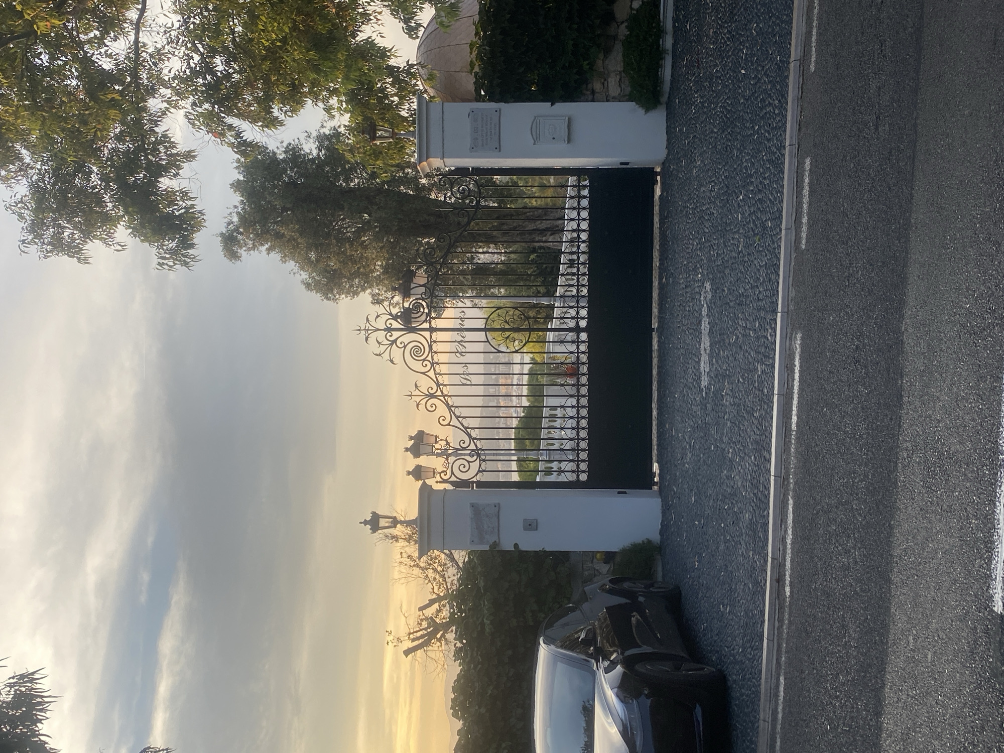The image captures a sunset scene in Antibes, France, featuring a large, ornate black metal gate leading to a property. The gate is flanked by two white pillars, each topped with a decorative lantern. A plaque is affixed to the left pillar. Beyond the gate, a glimpse of the sea and distant hills is visible. In the foreground, to the left of the gate, a portion of a black car is parked on a dark, pebbled driveway. Lush green foliage surrounds the entrance, and trees with autumn-colored leaves are visible in the background. The sky is filled with dramatic, cloudy formations illuminated by the warm hues of the setting sun.
