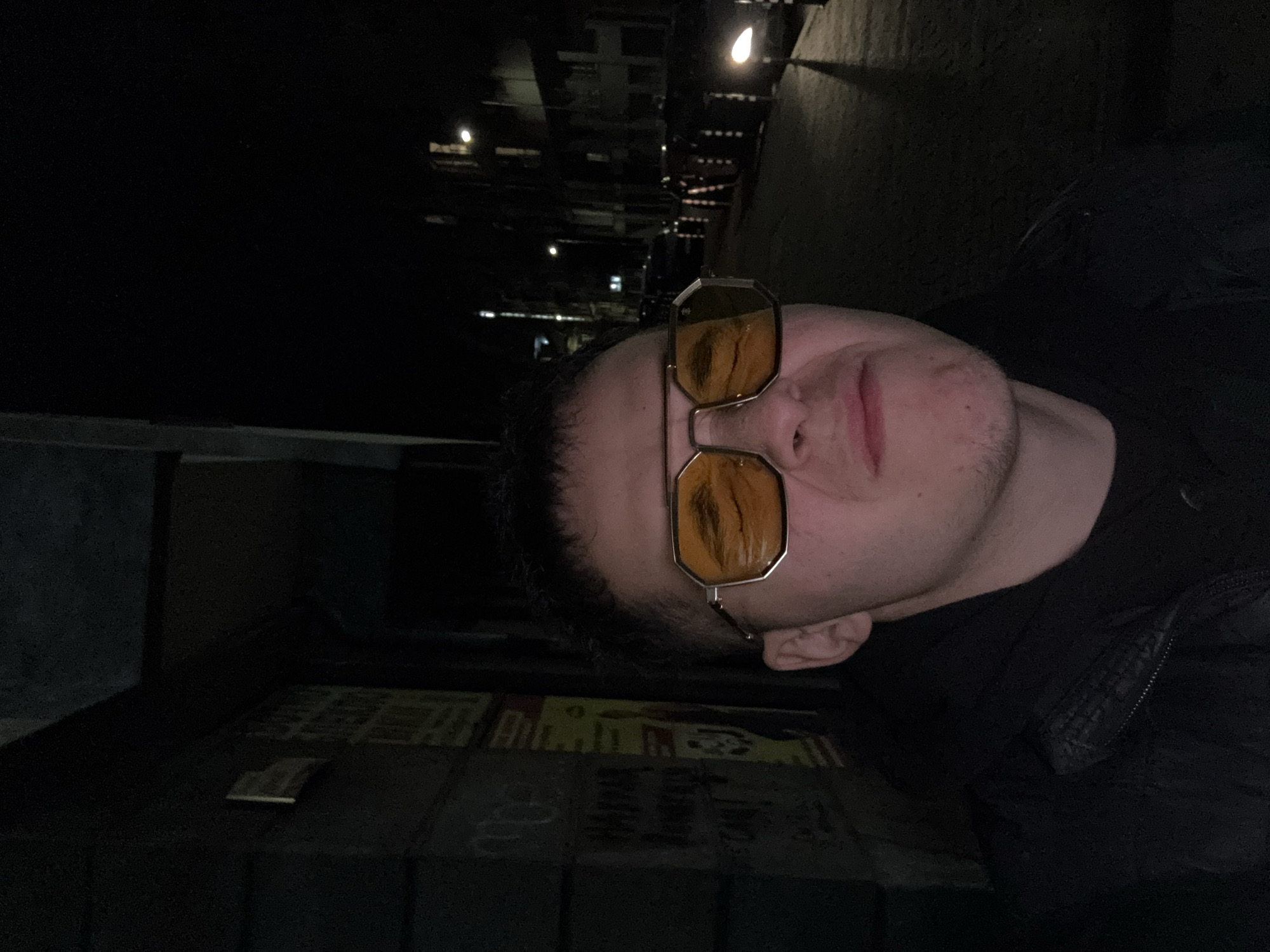 A nighttime close-up portrait of a person wearing yellow tinted glasses. The background shows buildings and streetlights in the city. The person is wearing a dark jacket. There are posters on a wall visible in the lower portion of the image. The picture is dark. Without more information and different angles, it is not possible to say the location in Sofia, Bulgaria.