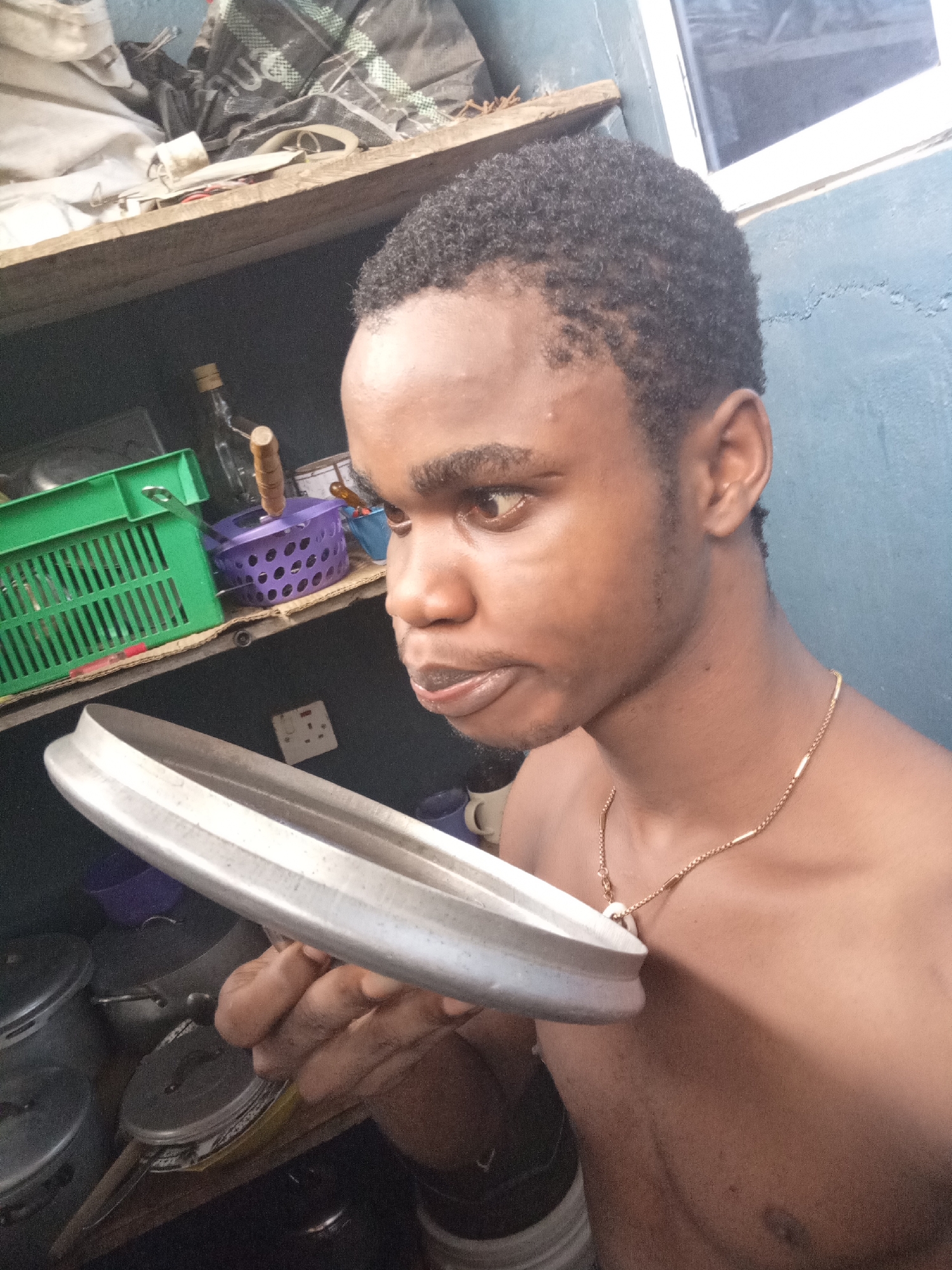 The image presents a candid portrait of a young man situated in an indoor environment, likely a kitchen or utility area in Owerri, Nigeria. The man, with a brown complexion, is shirtless, revealing a thin gold-colored chain with a small white pendant around his neck. He has short, dark, curly hair and a subtle amount of stubble on his chin and jawline. His eyes are wide and focused intently towards the right, beyond the frame, with his brows slightly furrowed and mouth partially open, conveying an expression of deep concentration, curiosity, or concern.
In his right hand, he holds a large, silvery, metallic ring-shaped object, which appears to be a component of a cooking pot, possibly a lid. The background reveals a domestic and functional space. A wooden shelf runs across the middle of the frame, supporting various items including a bright green plastic crate, a purple plastic basket, and a clear glass bottle. Below the shelf, several metal pots with lids are visible, suggesting cooking or storage implements. An electrical outlet is mounted on the muted blue-green wall behind the man's arm. A white-framed window or opening is partially visible in the upper right corner, providing an additional light source. The overall scene captures a moment of focused engagement within a lived-in setting. No discernible text is visible in the image.