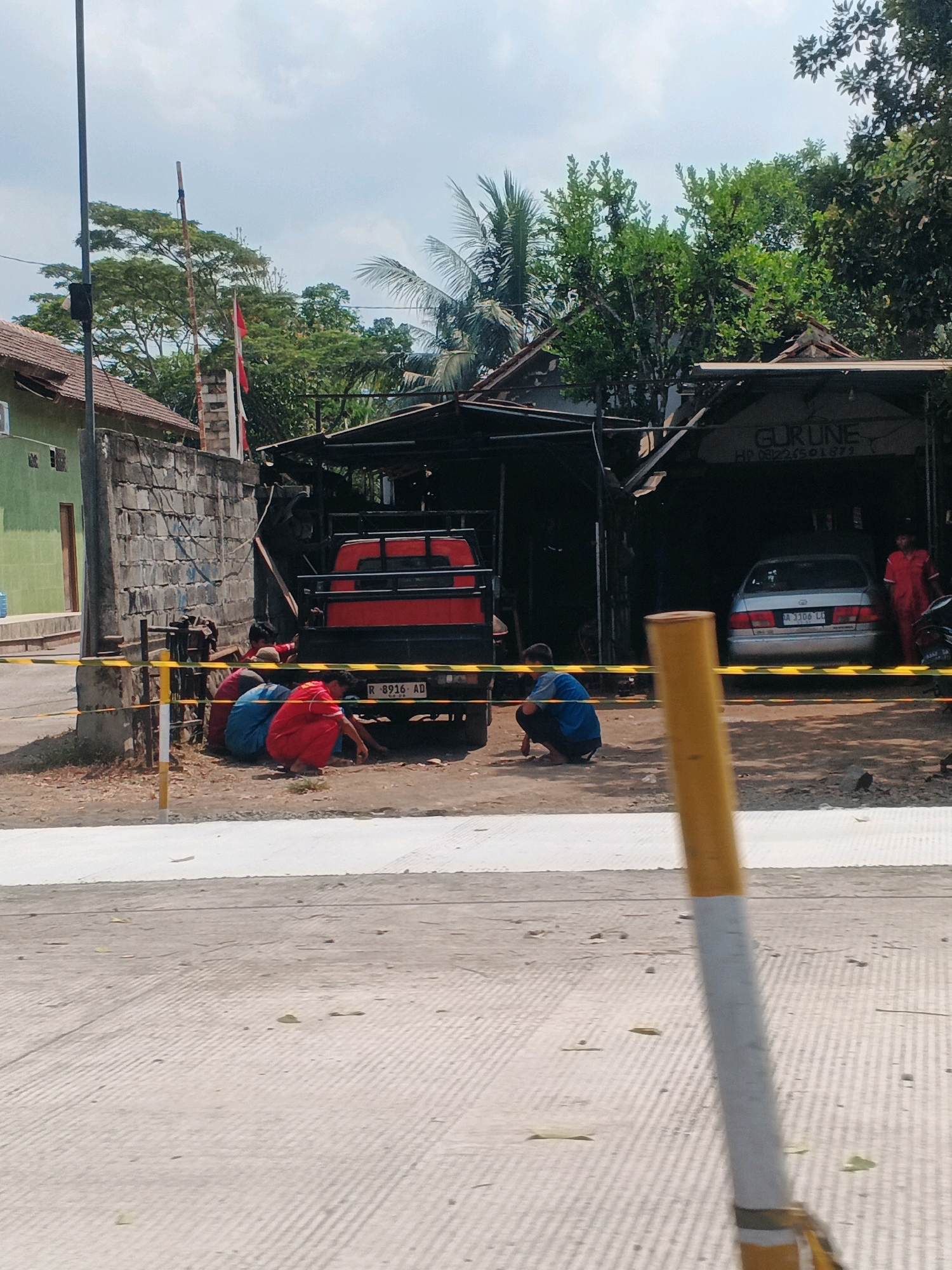 In Kedungpring, Indonesia, a street scene unfolds with a light blue sky overhead. A red pickup truck is parked in front of a structure with a corrugated metal roof, possibly a workshop or garage. Beside the truck, a silver sedan is also visible. Several individuals are present. Three people are crouched down near the rear of the pickup truck, two wearing red and one in blue. Another person in a blue shirt is squatting further to the right, near the entrance to the structure. A person in a red garment is standing near the silver sedan. A yellow and black striped barrier is set up across the foreground, partially obscuring the view of the scene. A tall, thin pole with a yellow top stands in the foreground. The ground appears to be a mix of dirt, gravel, and paved surfaces. Trees and foliage surround the area, with a building visible in the background to the left, painted green. The overall atmosphere suggests a casual or informal gathering or activity.