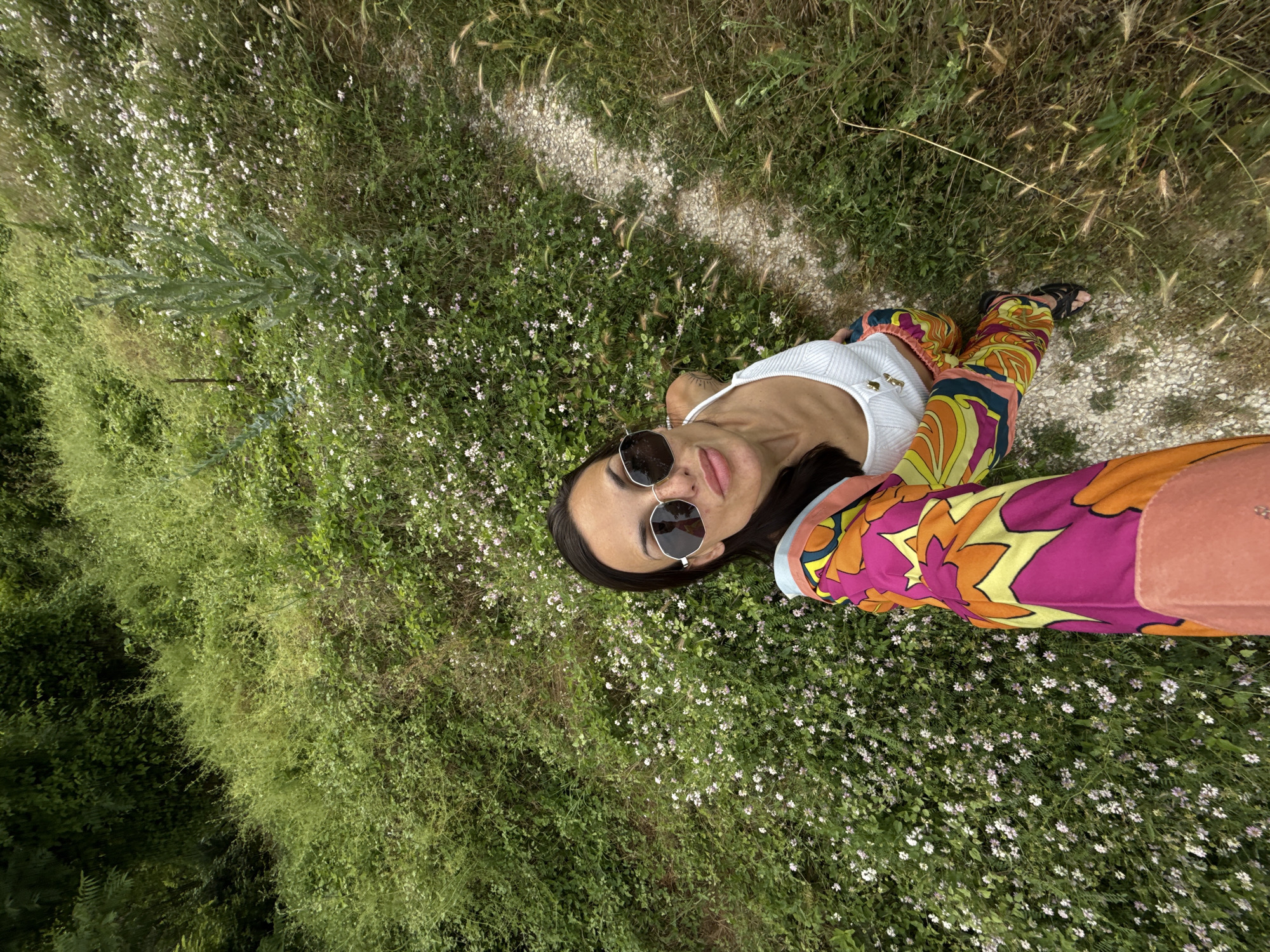 A woman is taking a selfie from a high angle in a grassy, outdoor location. She is wearing a white crop top, colorful patterned pants, and sunglasses. The background is a hillside covered in lush green grass and small white flowers, with a dirt path winding to the right. The woman is smiling and looking at the camera. There is no indication of the city of Pleven, Bulgaria.