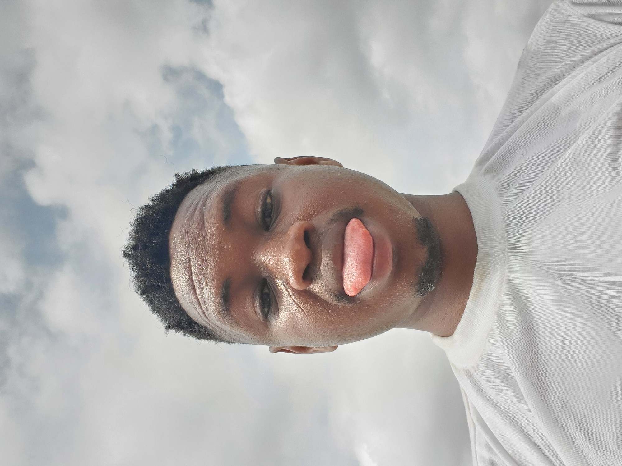 A close-up, slightly angled upward selfie shows a Black male with short, dark, textured hair and a small beard, sticking his tongue out. He wears a white t-shirt. The background is a cloudy sky. No specific location in Mararaba, Nigeria, can be determined from the image.