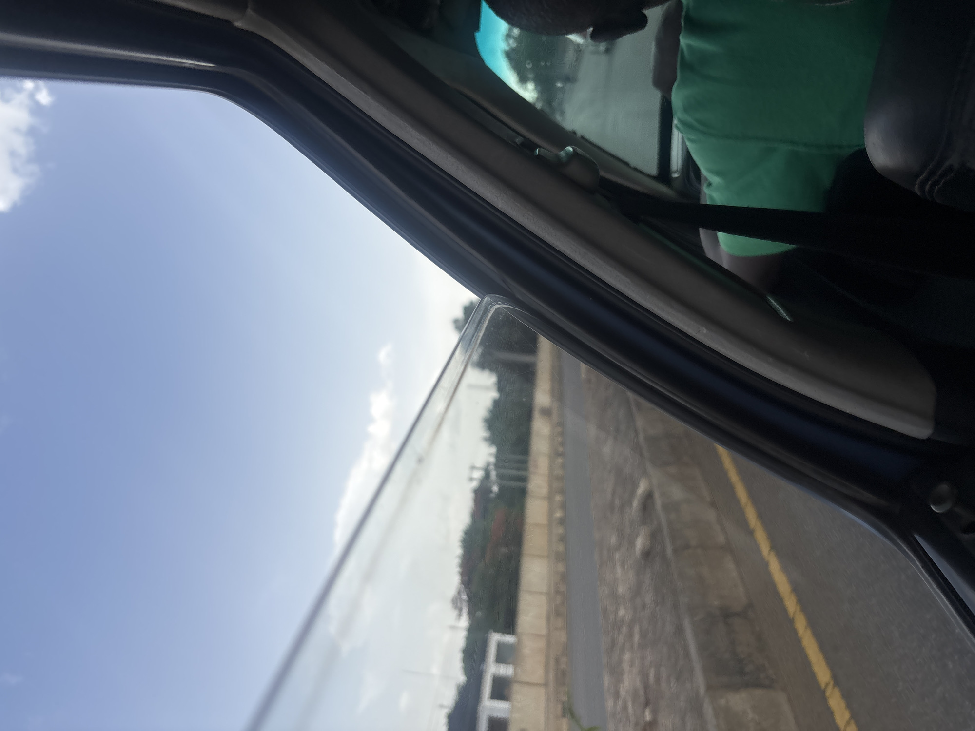 A person wearing a green t-shirt is visible from the back, seated in the passenger seat of a vehicle. The view is through the open window, looking outwards. Outside the vehicle, a road with a yellow line marking is visible, bordered by a concrete barrier. Beyond the barrier, there is a building with white walls and a flat roof, and further in the distance, trees and utility poles can be seen. The sky is bright blue with some scattered clouds. The scene appears to be in a city environment, potentially in Jos, Nigeria, given the characteristic architecture and landscape.