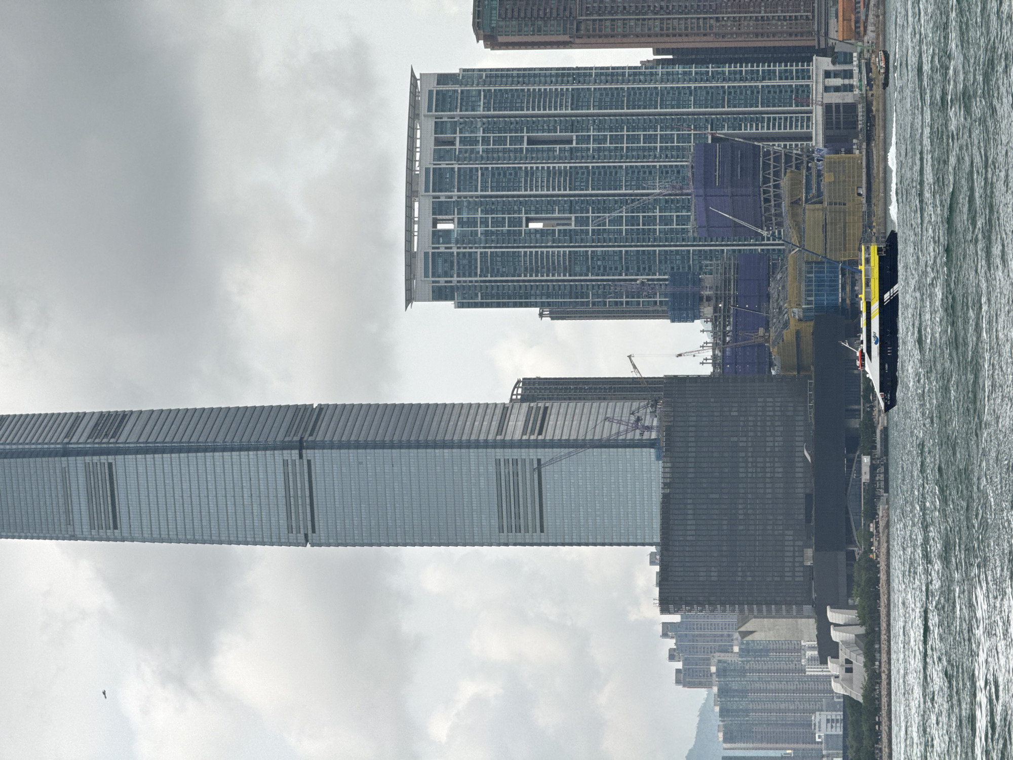 The image shows a cityscape in Hong Kong, China, featuring several tall buildings along a waterfront. The sky is overcast. The International Commerce Centre (ICC) is visible on the left, and other high-rise buildings are in the background. There is a body of water on the right side of the image. Construction is underway near some of the buildings, with visible cranes and scaffolding. A boat is near the waterfront.