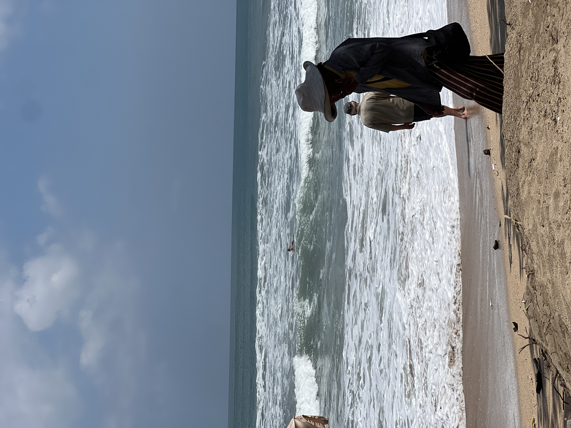 This image captures a scene at the beach in Kuta, Indonesia. In the foreground, a person wearing a wide-brimmed hat and a striped outfit is partially visible, with their hand near their face. Further back, another individual in a light-colored shirt and shorts stands with their back to the camera, facing the ocean. A surfer is visible in the distance, riding a wave in the turquoise water. The ocean is active with white-capped waves breaking near the shore, and the sky above is a mix of blue with scattered clouds. The beach itself is sandy, with some debris and vegetation visible along the waterline.