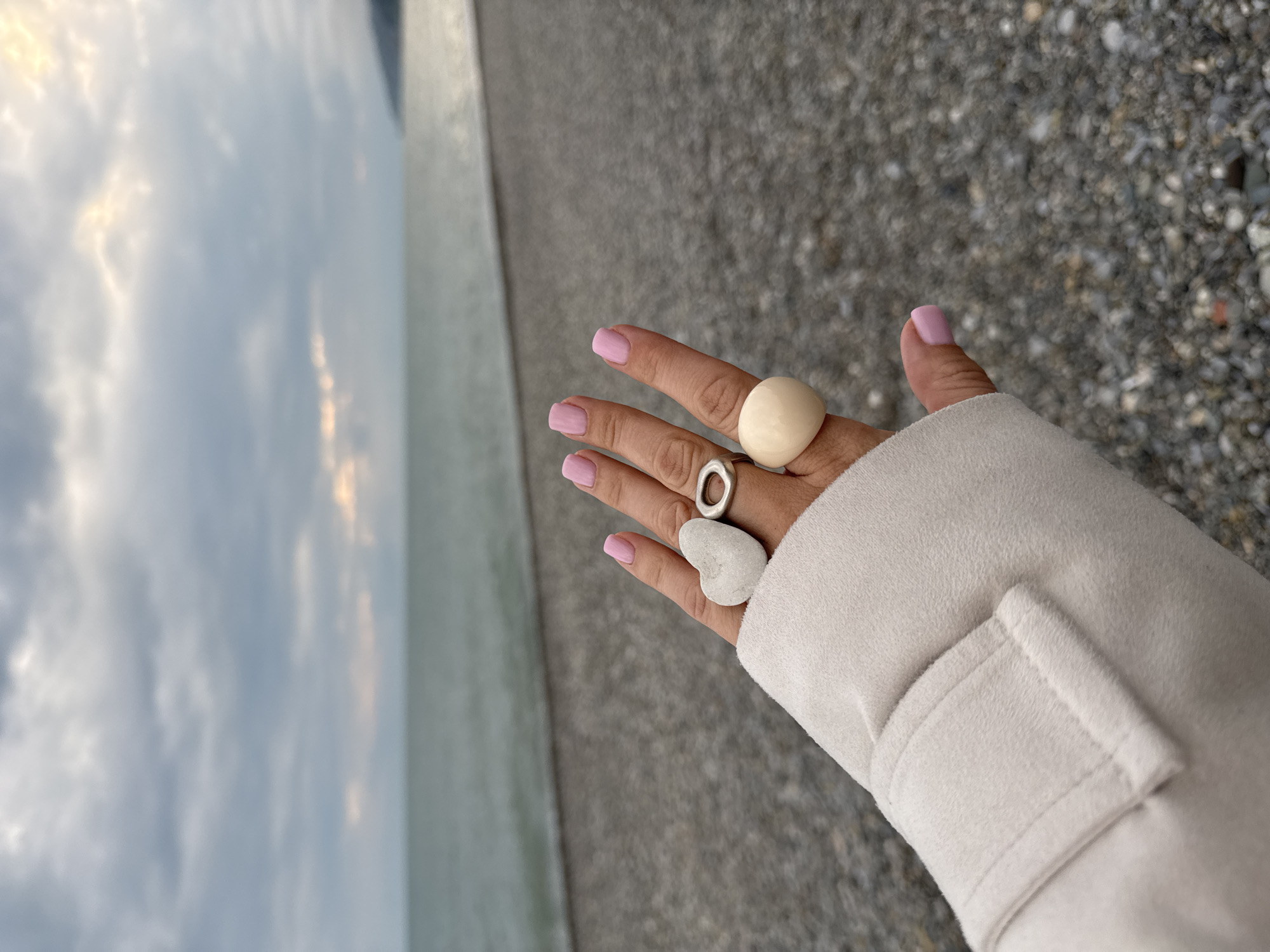 A hand, adorned with two rings and holding a heart-shaped stone, is presented against the backdrop of a beach in Sochi, Russia. The hand, with pink nail polish, wears a large, round, cream-colored ring on the middle finger and a silver ring with a hollow center on the ring finger. A small, white, heart-shaped stone rests on the back of the hand. The hand is partially covered by a light-colored coat sleeve. The beach consists of small stones and pebbles, and the sea is visible in the distance under a cloudy sky.