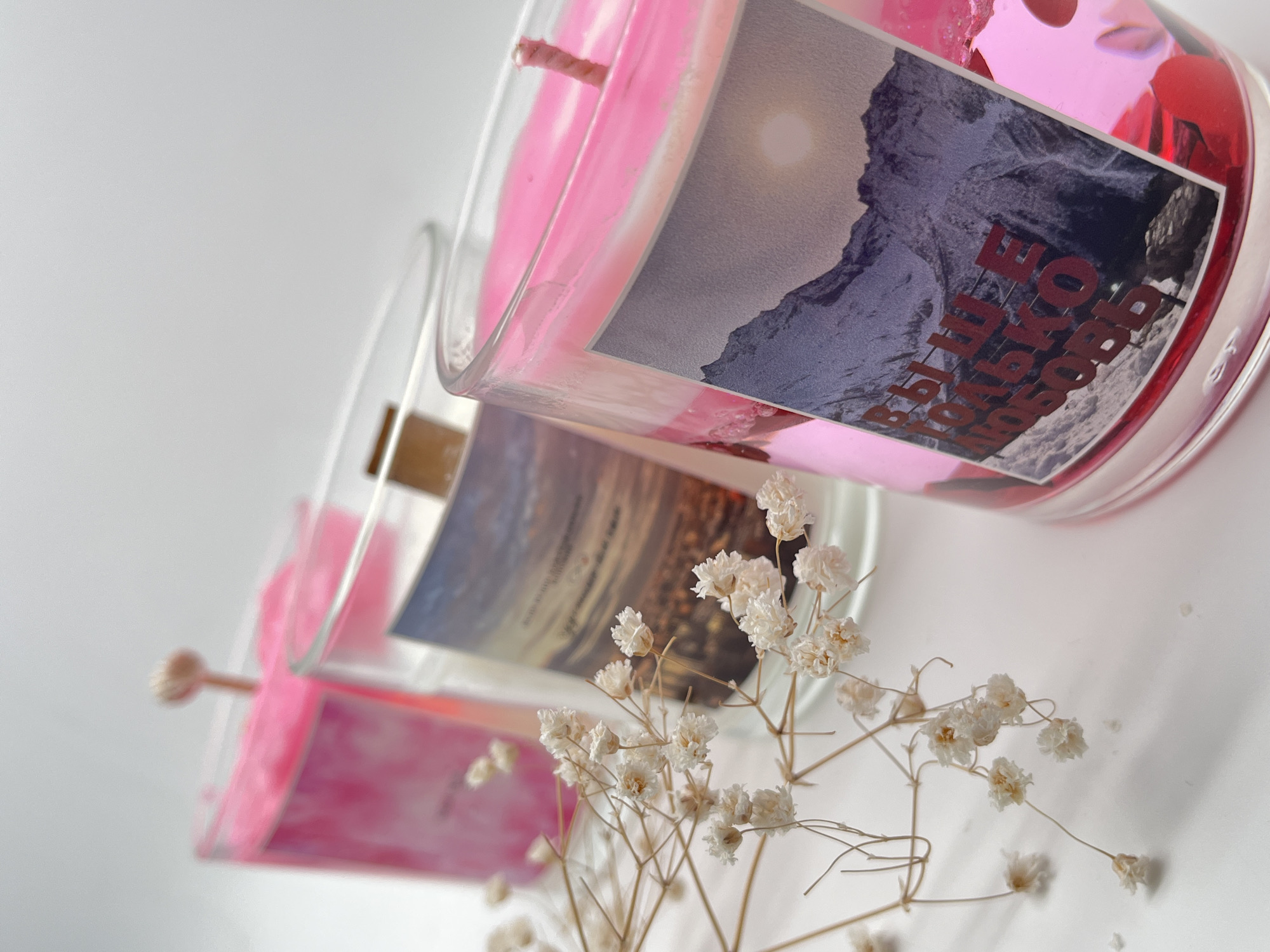 An overhead, slightly angled shot displays two decorative pink candles in clear glass jars, arranged diagonally against a plain white background. In the foreground, a prominent candle features bright pink wax and is adorned with small, reflective, pink heart-shaped confetti or flakes. A label on this candle shows a photographic image of a rugged mountain range under a luminous sun or moon. Overlaid on this mountain image is Cyrillic text in dark red, partially legible as "ВЫШЕ ТОЛЬКО ЛЮБОВЬ" (VYSHE TOL'KO LYUBOV', meaning "Only love is higher"). The text is oriented vertically upwards.
Behind it, a second, slightly blurred candle with pink wax also sits in a clear glass jar, featuring a label depicting a serene sunset or sunrise scene over what appears to be water or a distant horizon. The text on this second candle is indistinct due to the blur.
Scattered across the foreground and midground are delicate sprigs of dried white or cream baby's breath (Gypsophila) flowers, adding a soft, natural element to the composition. The overall lighting is bright and even, highlighting the vibrant pink of the candles and the textures of the labels and flowers. The Cyrillic text on the candle suggests a Russian context, aligning with the provided location context of Perm, Russia.