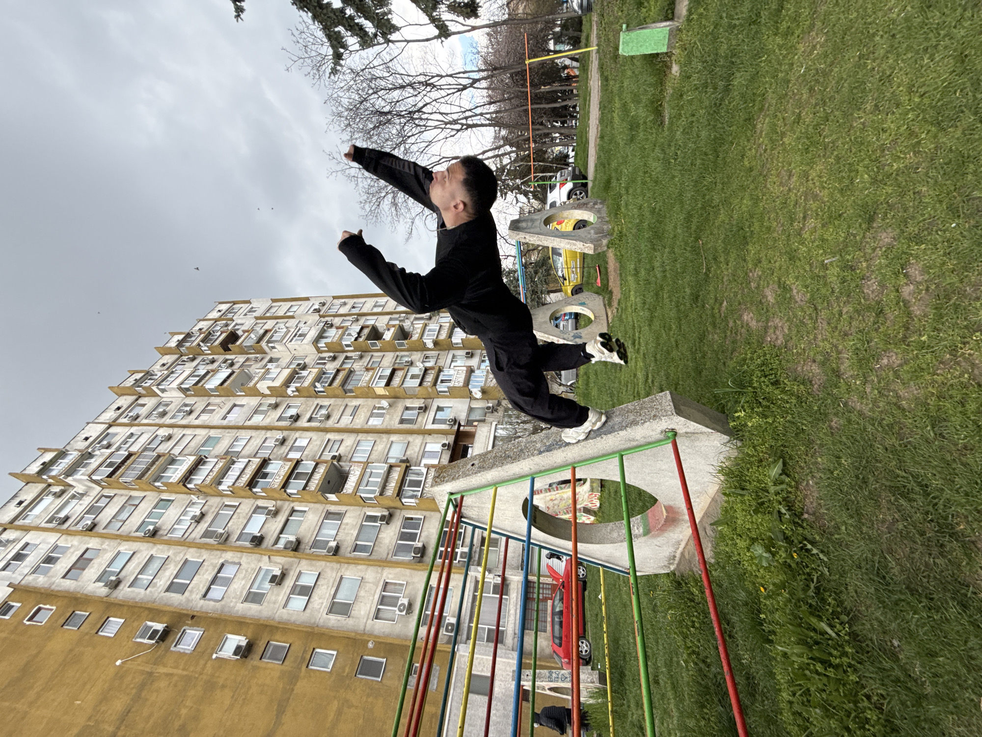 The image captures a young man in a dynamic pose atop a concrete playground structure. He is dressed in black clothing and white sneakers, with his arms outstretched as if in flight. The background consists of a tall, multi-story residential building with numerous windows and air conditioning units. Adjacent to the playground is a green grassy area with trees and other playground equipment. The colorful metal bars of the playground structure add a vibrant contrast to the scene.
The location is Shumen, Bulgaria.